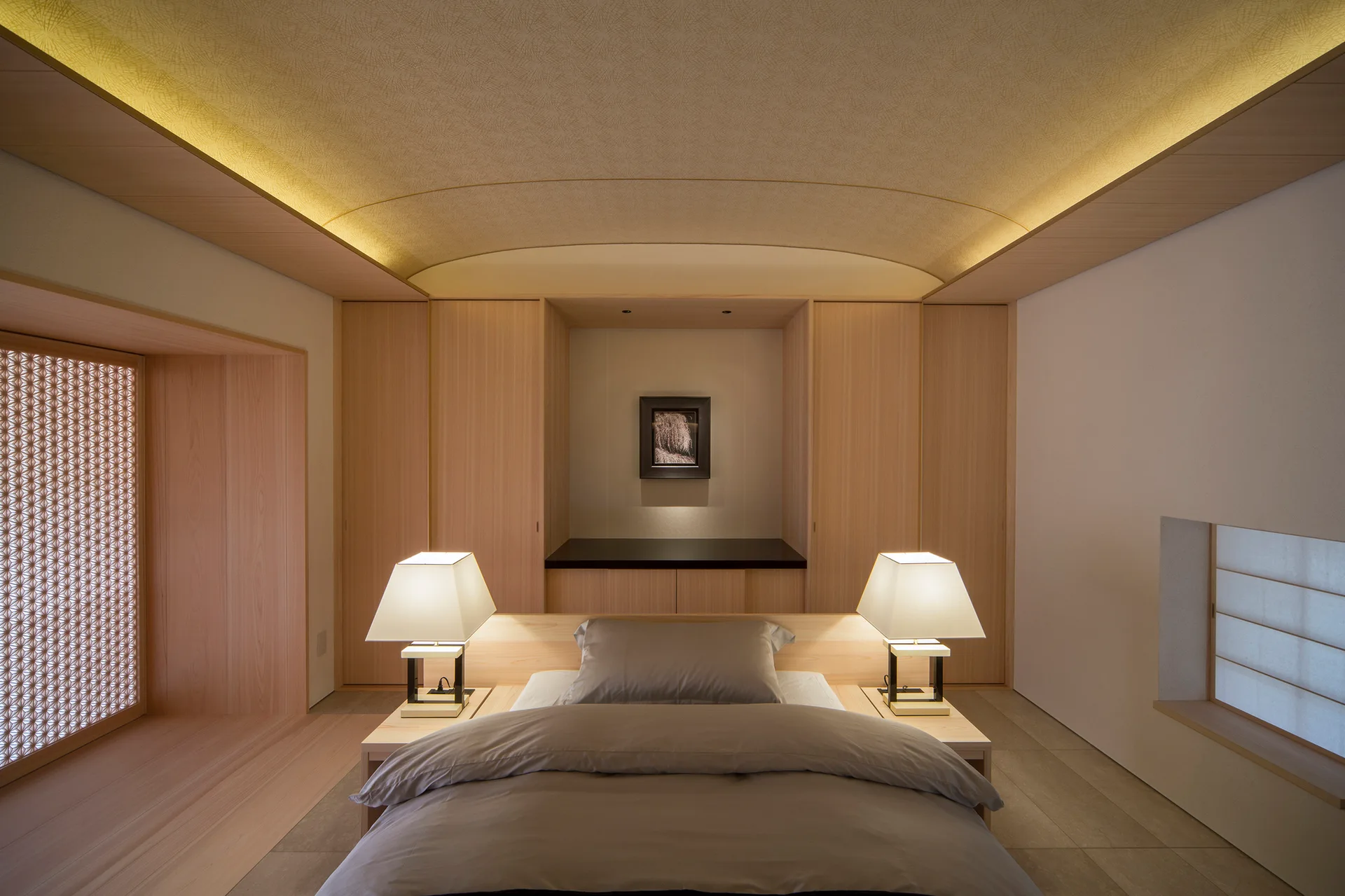 A minimalist Japanese style bedroom featuring a central bed flanked by twin table lamps beneath a curved wood ceiling with warm indirect cove lighting.