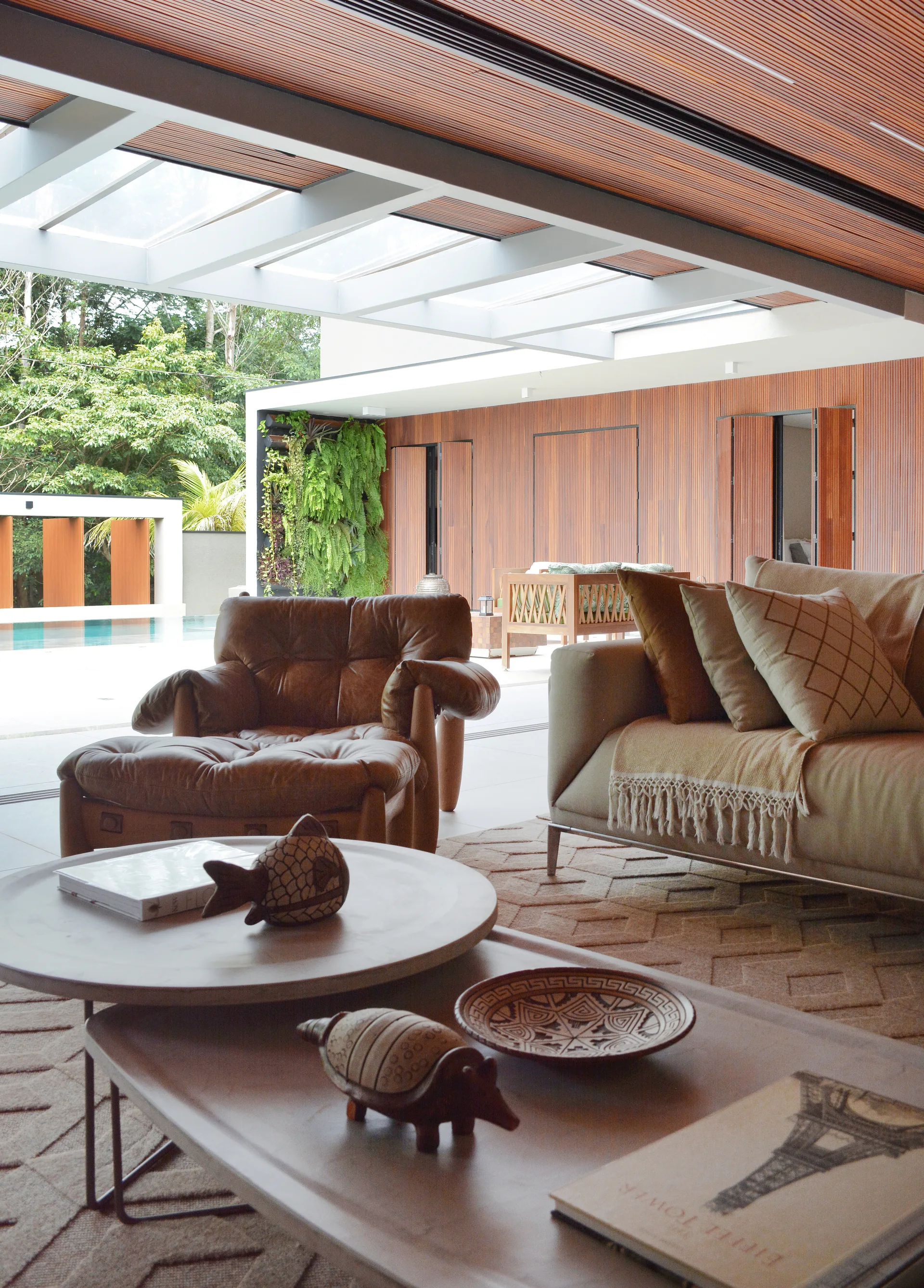 A covered patio features a leather armchair and a tan sofa arranged under a slatted wood ceiling with skylights, looking out toward a swimming pool.