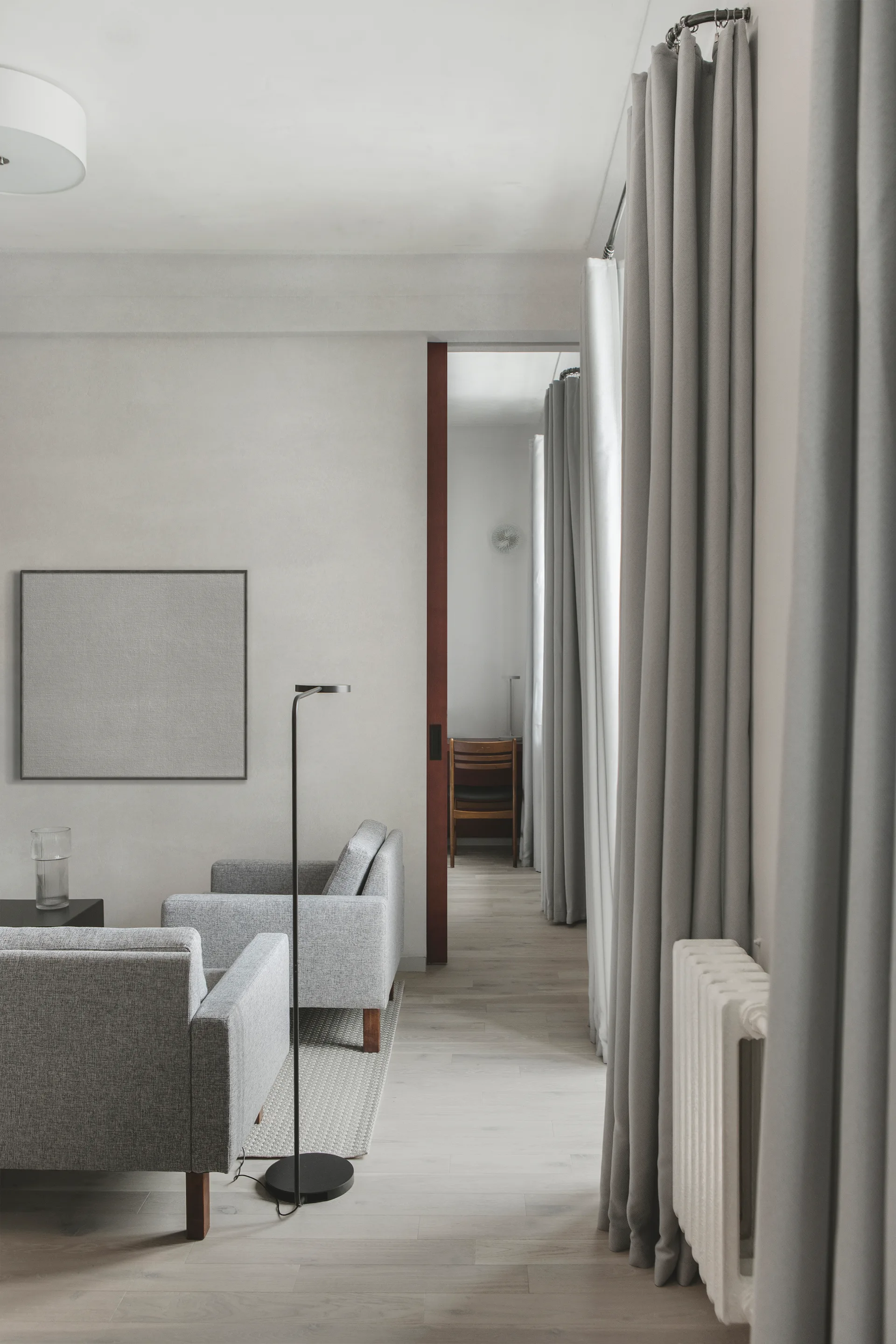 A minimalist living room features grey upholstered armchairs, a slender black floor lamp, and tall grey curtains next to a doorway.
