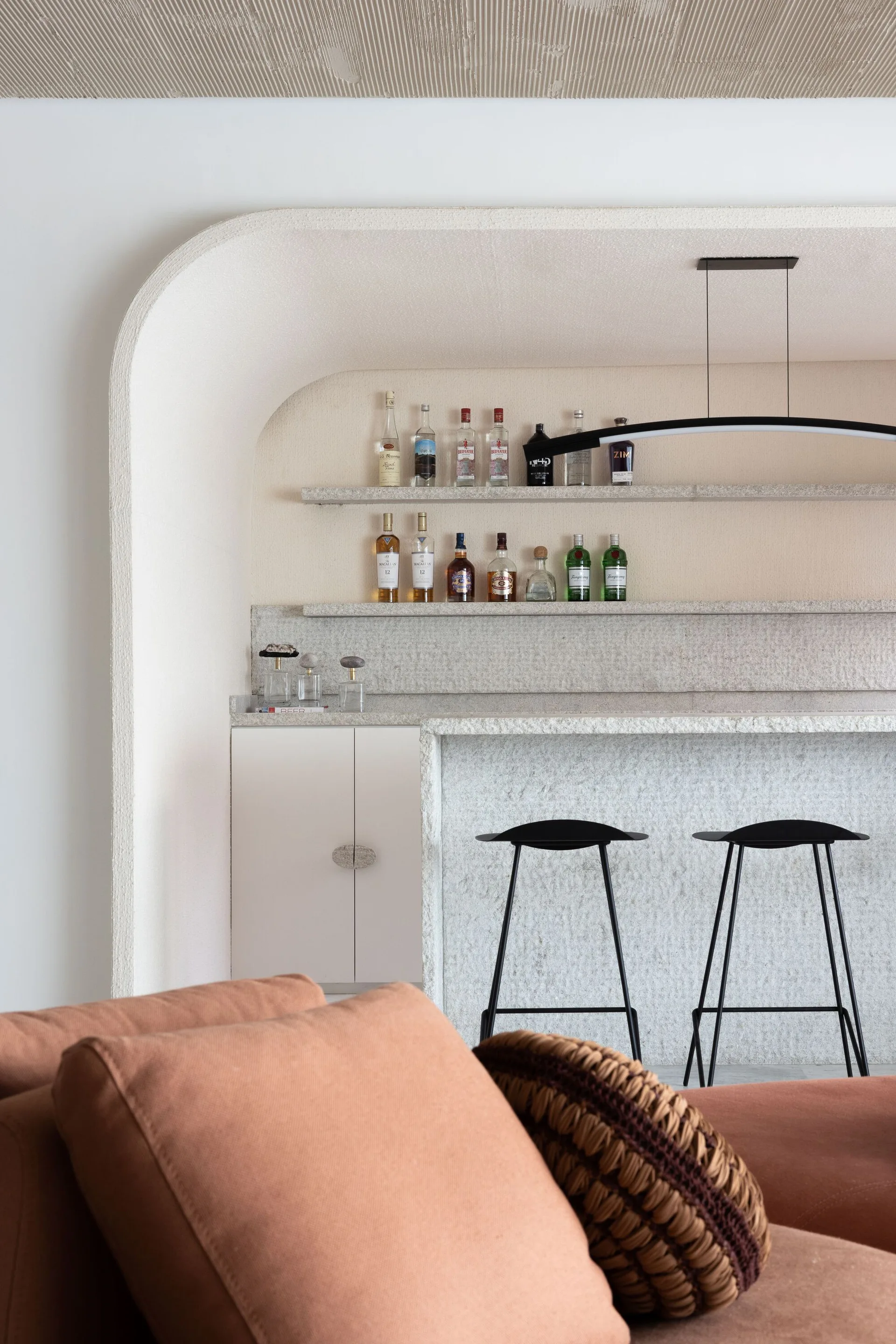 A minimalist home bar featuring a textured stone archway with recessed shelves, a matching stone counter, and two black metal bar stools.