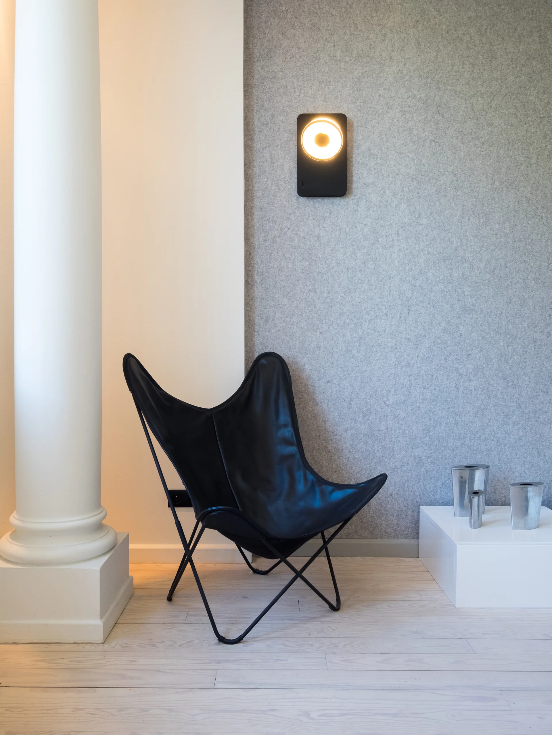 A black leather butterfly chair sits on light wood flooring between a white column and a textured grey wall with a circular wall light.