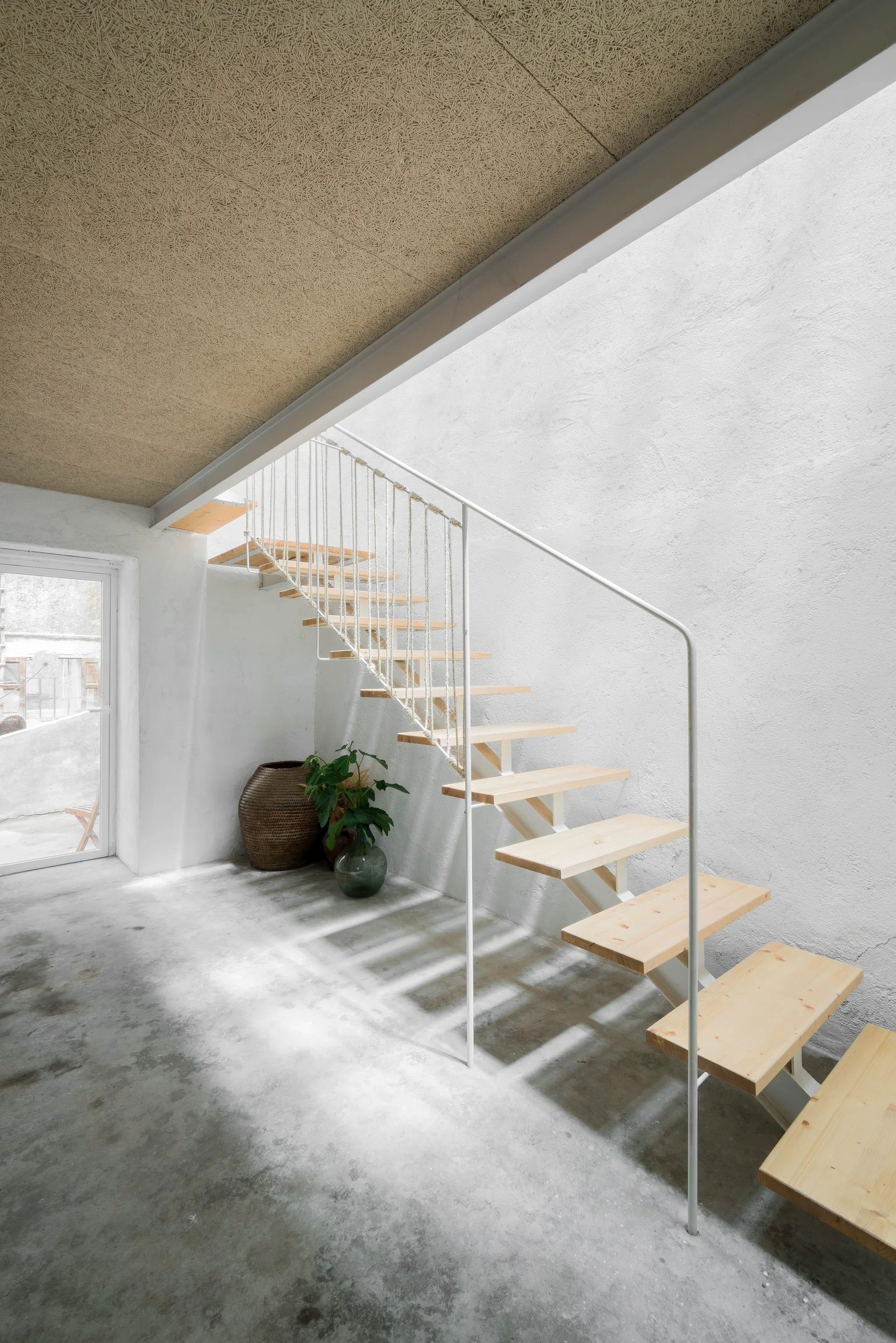 A minimalist open-riser wooden staircase features vertical rope railings next to a textured white wall and concrete floor.