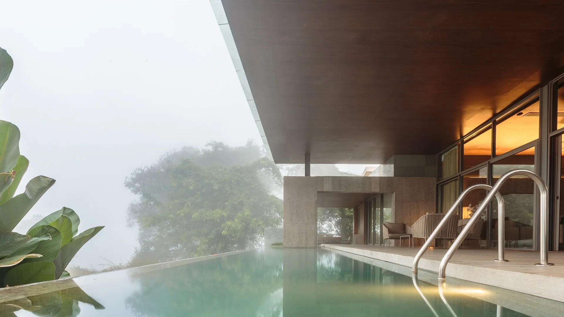 An infinity edge pool sits next to a covered stone patio with lounge chairs under a dark wood ceiling overlooking a misty forest.