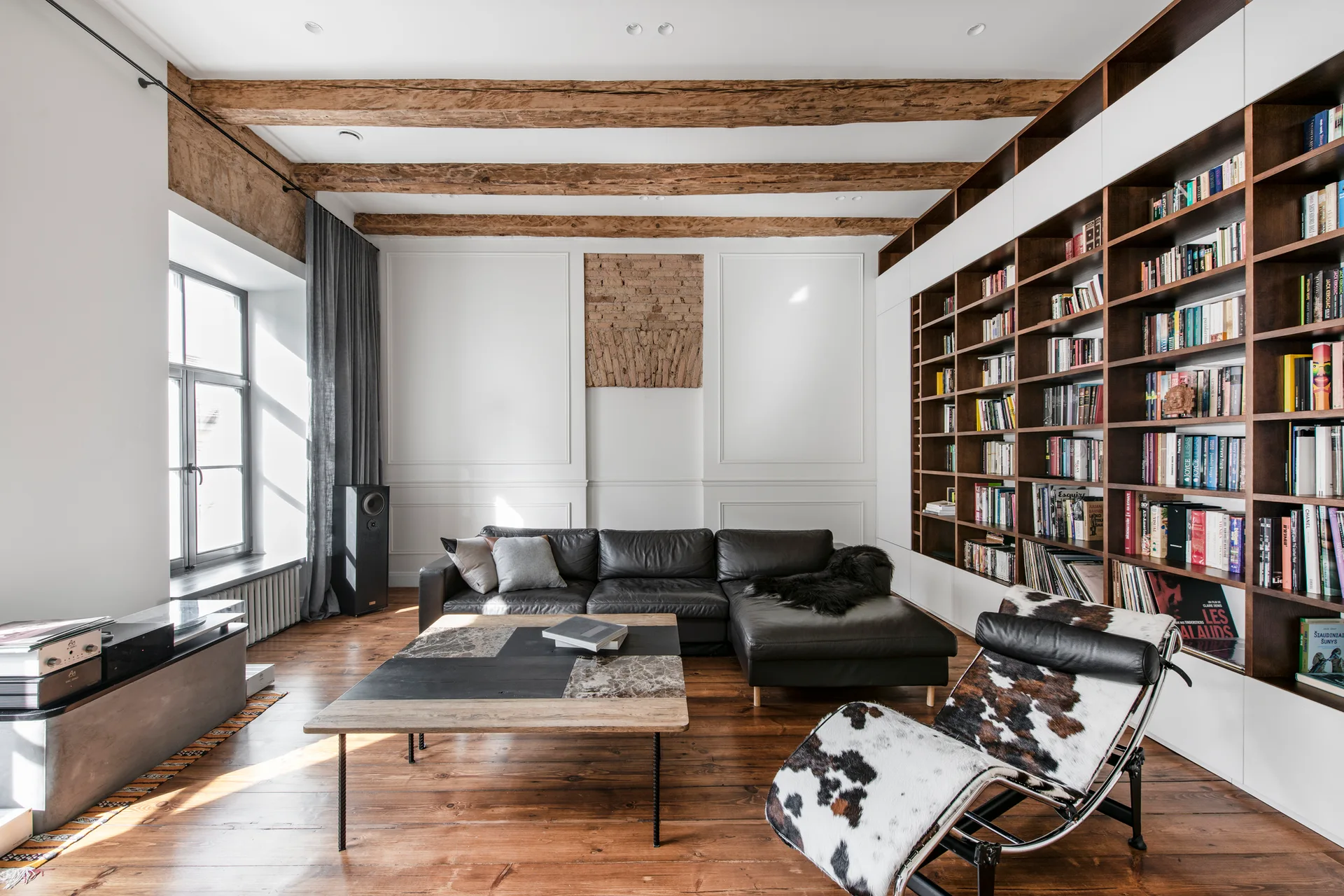A spacious living room features exposed wood ceiling beams, a large built-in bookshelf filled with books, a black leather sectional, and a cowhide lounge chair.