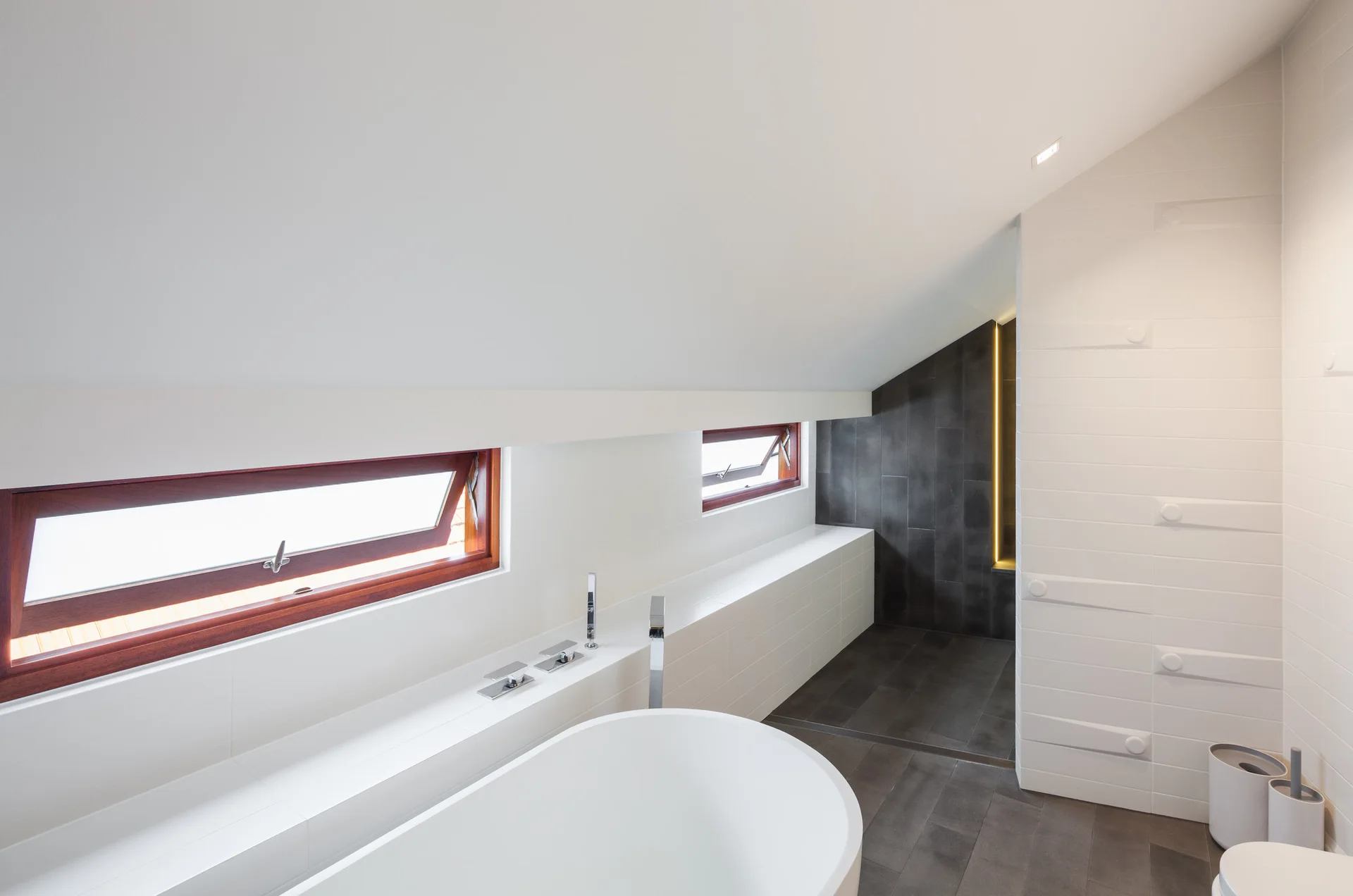 A minimalist attic bathroom featuring a white freestanding bathtub beneath a sloped ceiling with wood-framed windows and dark grey floor tiles.