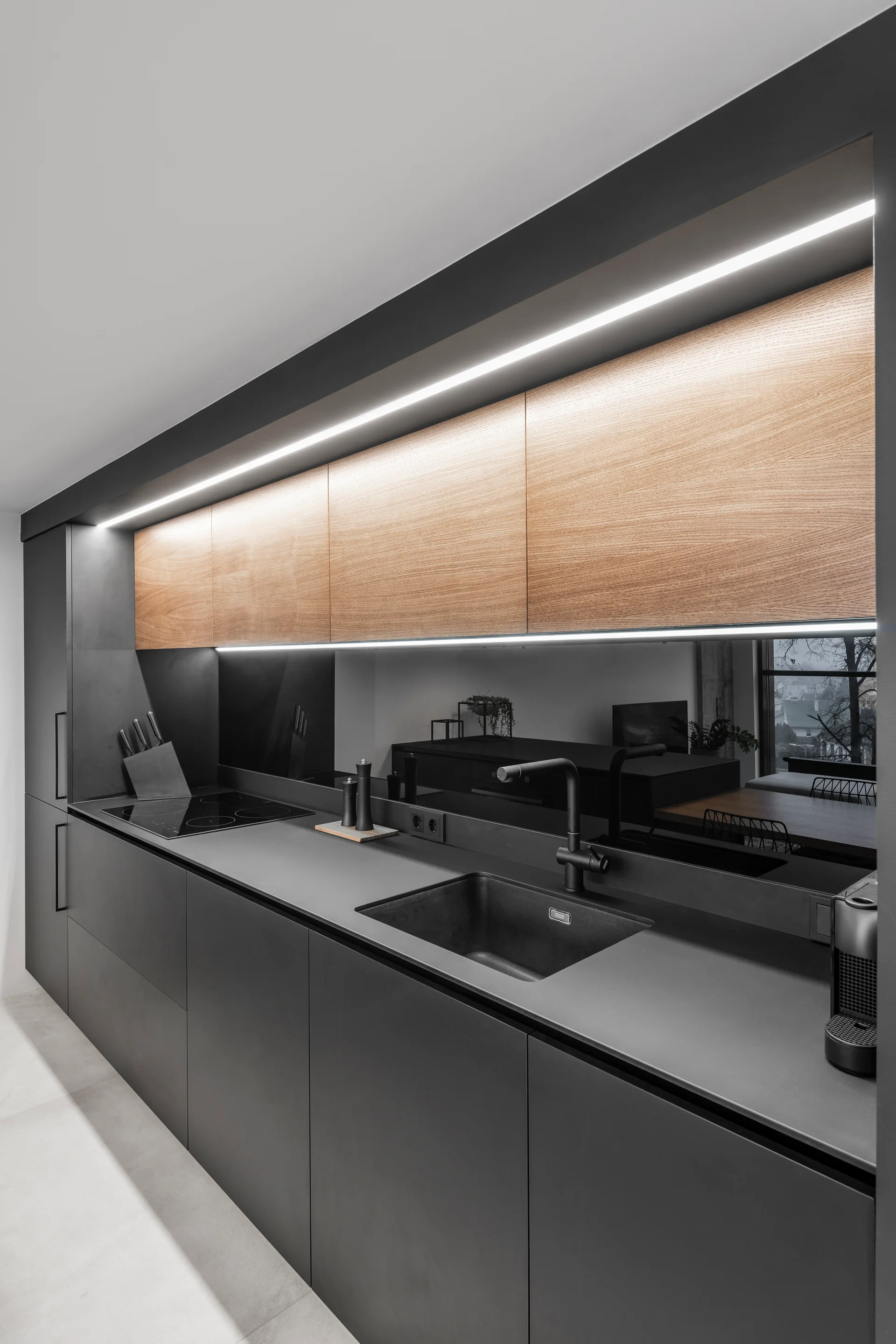 A modern kitchen featuring matte black base cabinets, warm wood upper cabinets, an integrated black sink, and a dark mirrored backsplash with LED lighting.