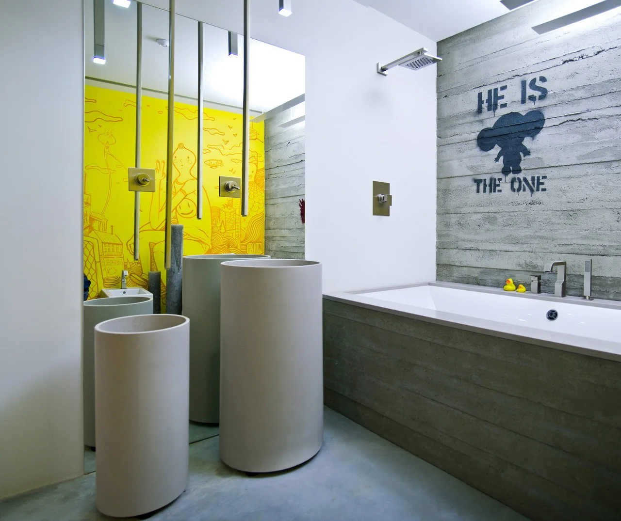 A modern bathroom with a concrete bathtub, cylindrical pedestal sinks, and a large mirror reflecting a yellow wall mural.