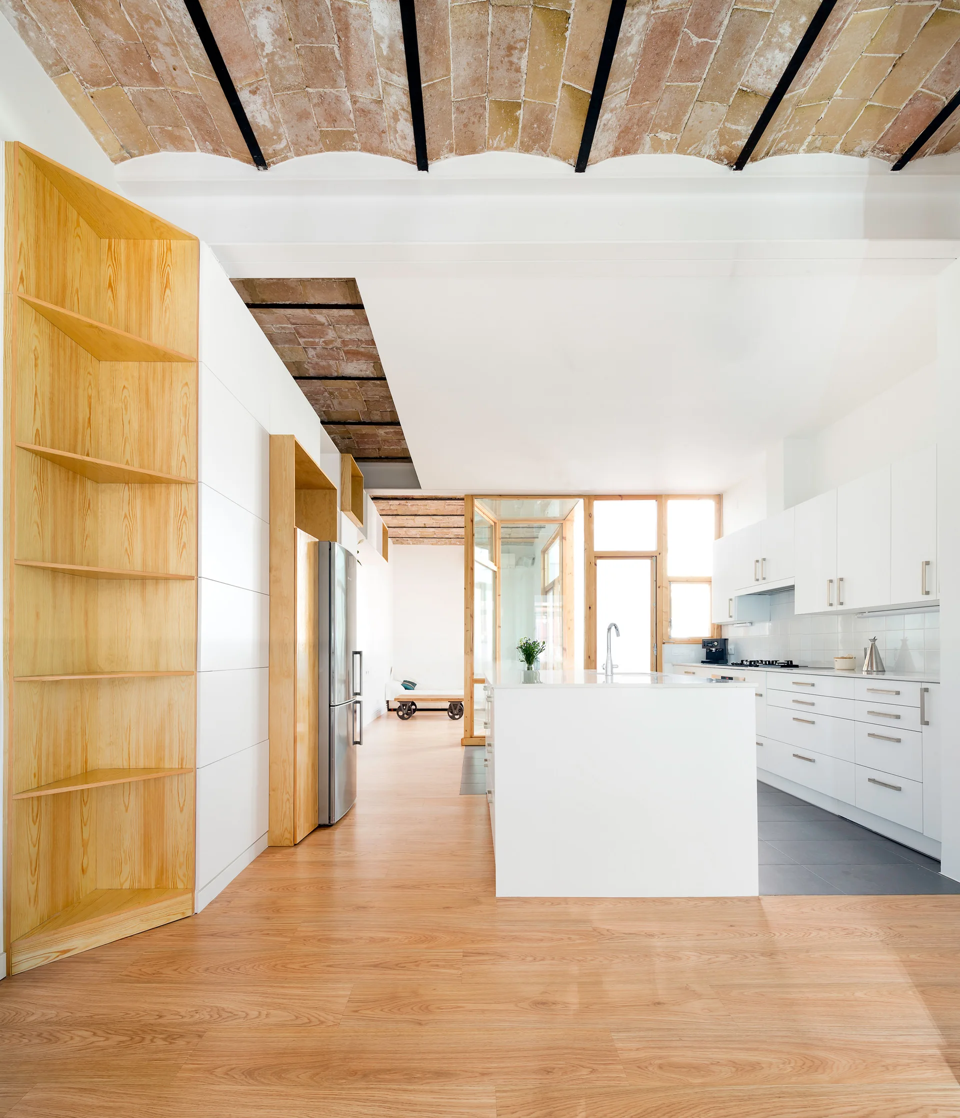A modern white kitchen features a vaulted brick ceiling, light wood flooring, a matching tall wood shelving unit, and a central white island.