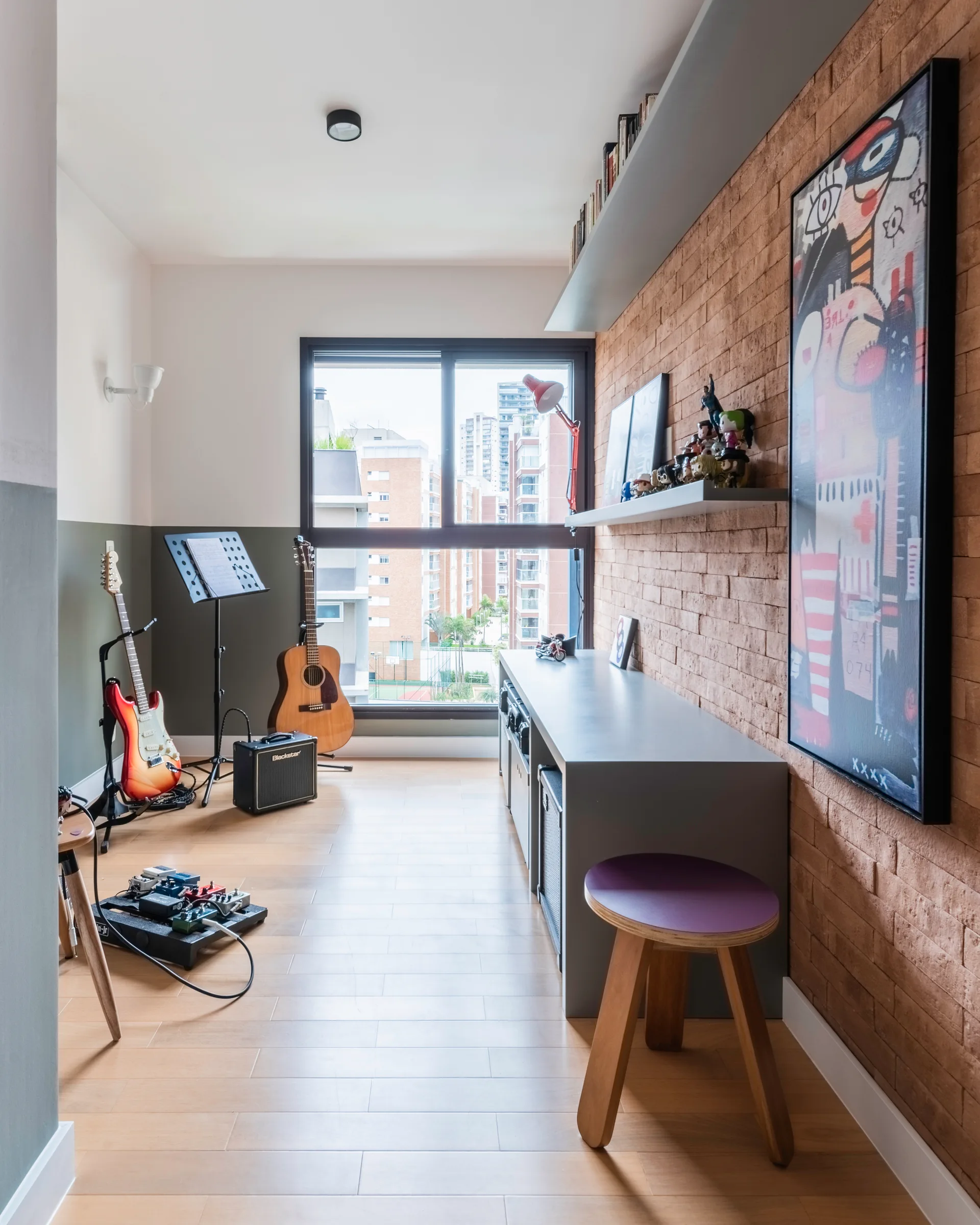 A home music studio features a textured brick wall with a low grey desk, floating shelves, and guitars on stands by a large window.