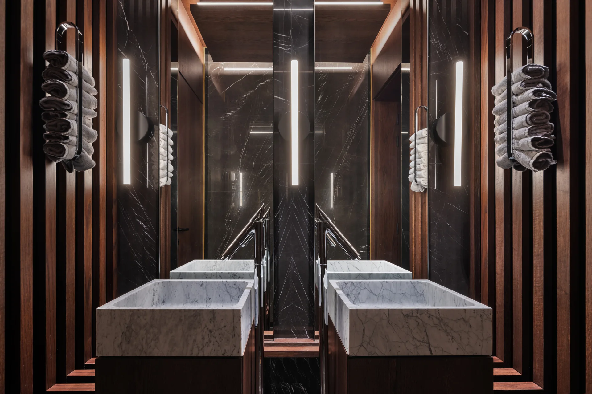 Symmetrical bathroom vanity with two block marble sinks, dark wood slat walls, and vertical tube lights integrated into large mirrors.