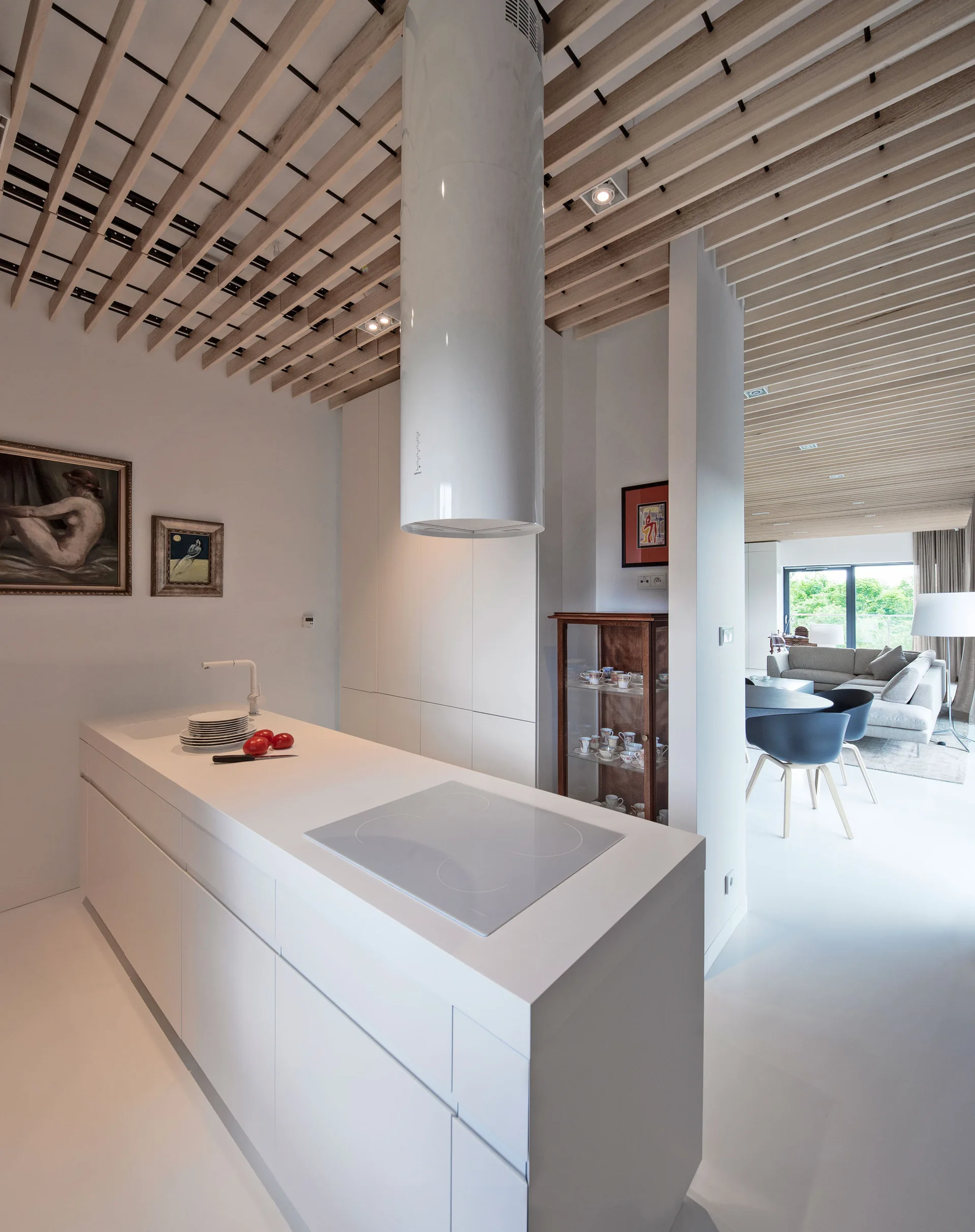 A modern white kitchen featuring a central island with a cylindrical hood under a slatted wood ceiling, opening to a distant living room.