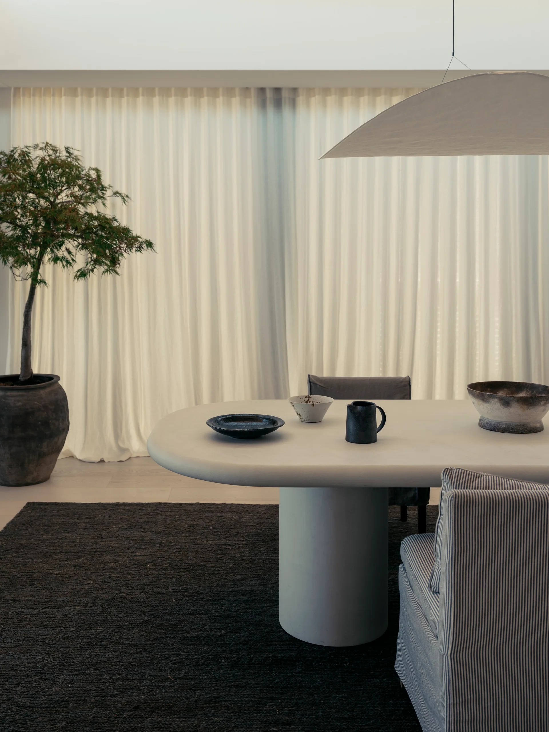 An organic modern dining room featuring a white oval pedestal table, pinstriped upholstered chairs, and a large potted tree against sheer white drapes.