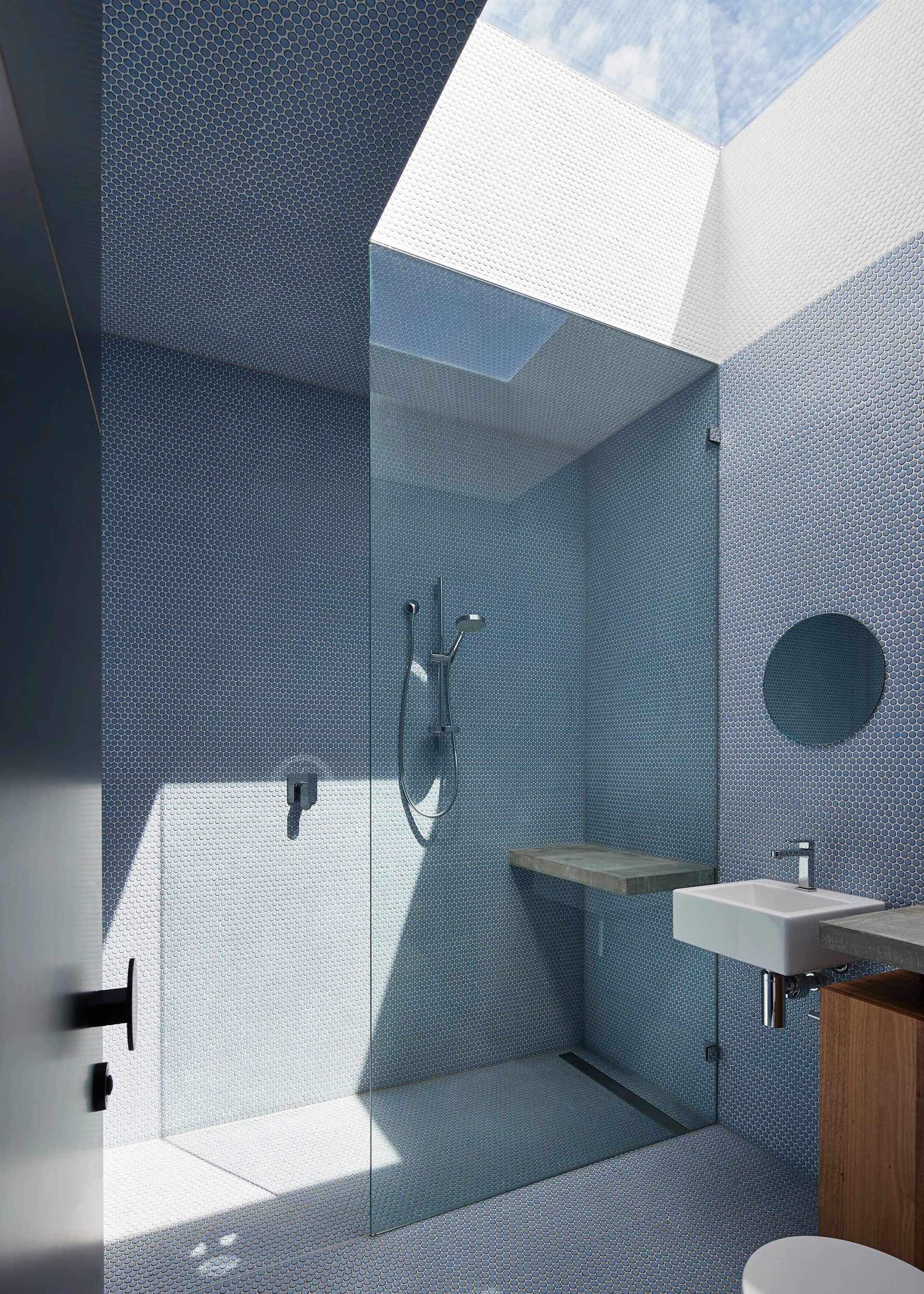 A modern bathroom features blue penny-round tile walls and floors, a glass-enclosed shower, a floating vanity, and a large skylight overhead.