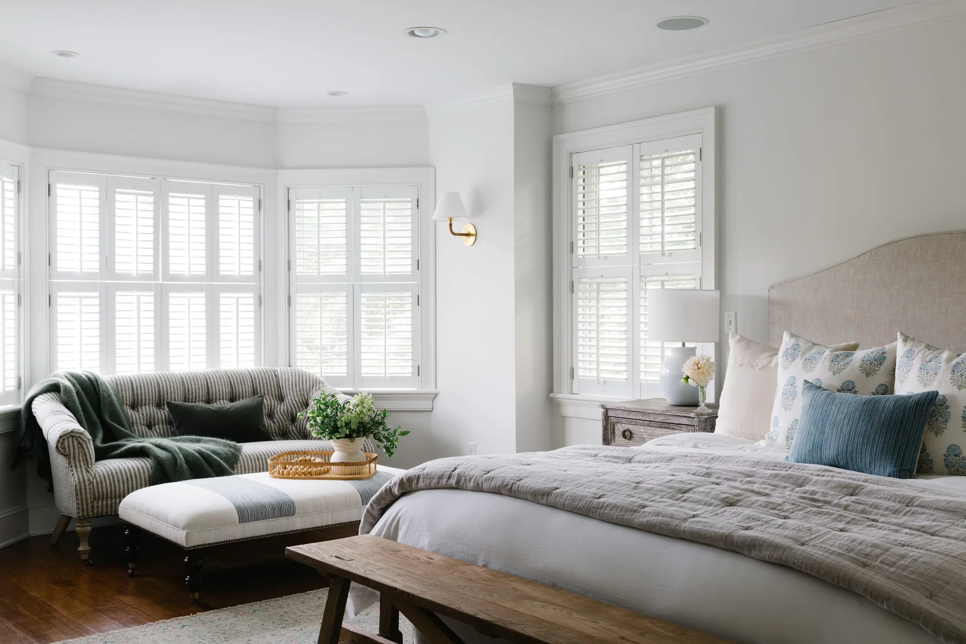 A bright bedroom featuring an upholstered bed with patterned pillows, a wooden foot bench, and a bay window seating area with a striped sofa.