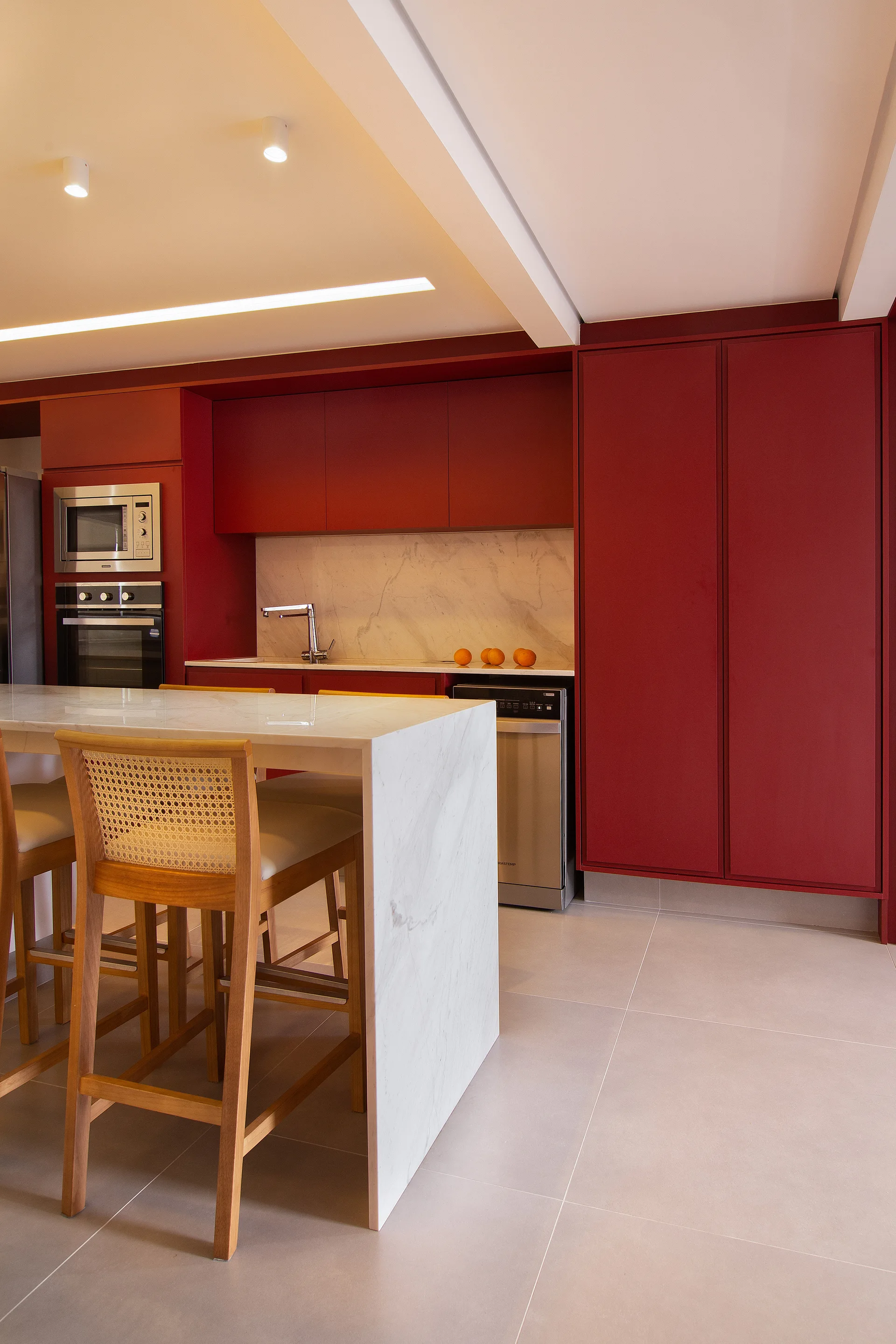 A modern kitchen featuring matte red cabinetry, a white marble waterfall island with wooden bar stools, and integrated stainless steel appliances.