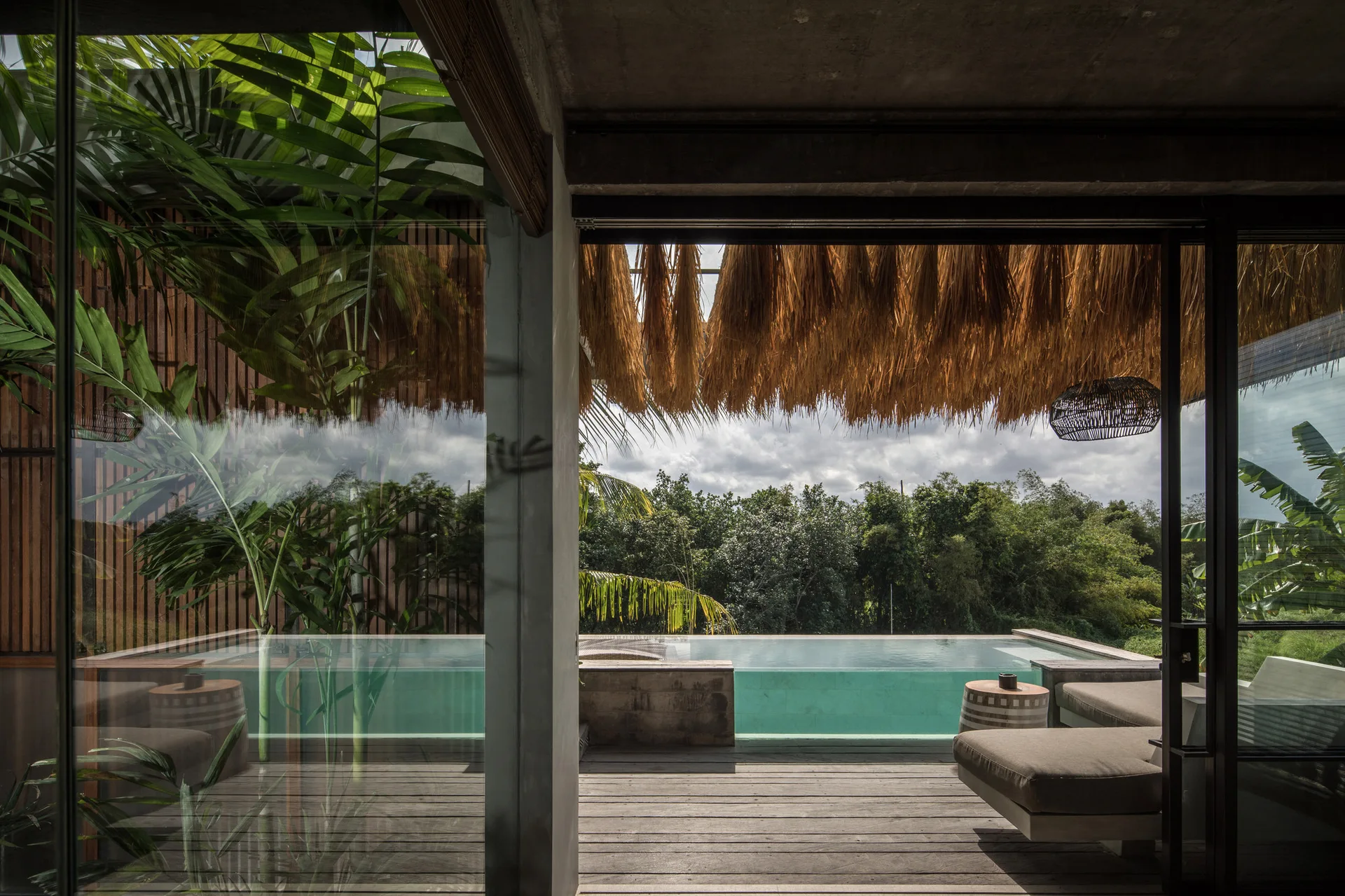 A view from a concrete interior looking out onto a wooden deck with lounge seating and an infinity pool under a thatched roof structure.
