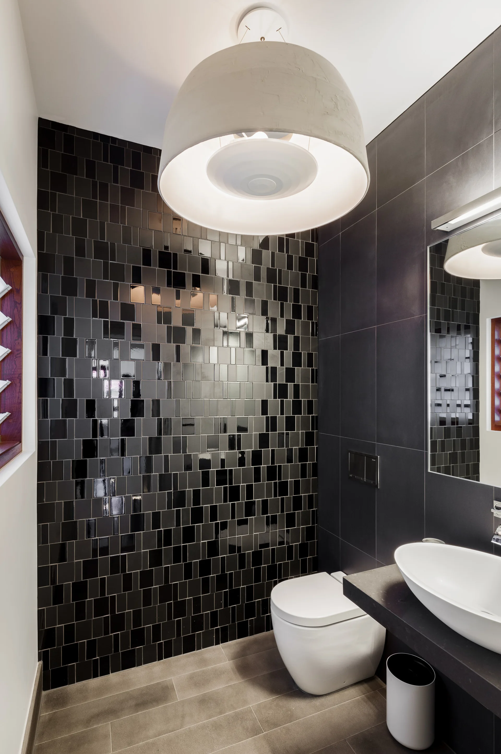A modern powder room featuring a glossy black mosaic tile accent wall, a white wall-hung toilet, and a large round white pendant light.