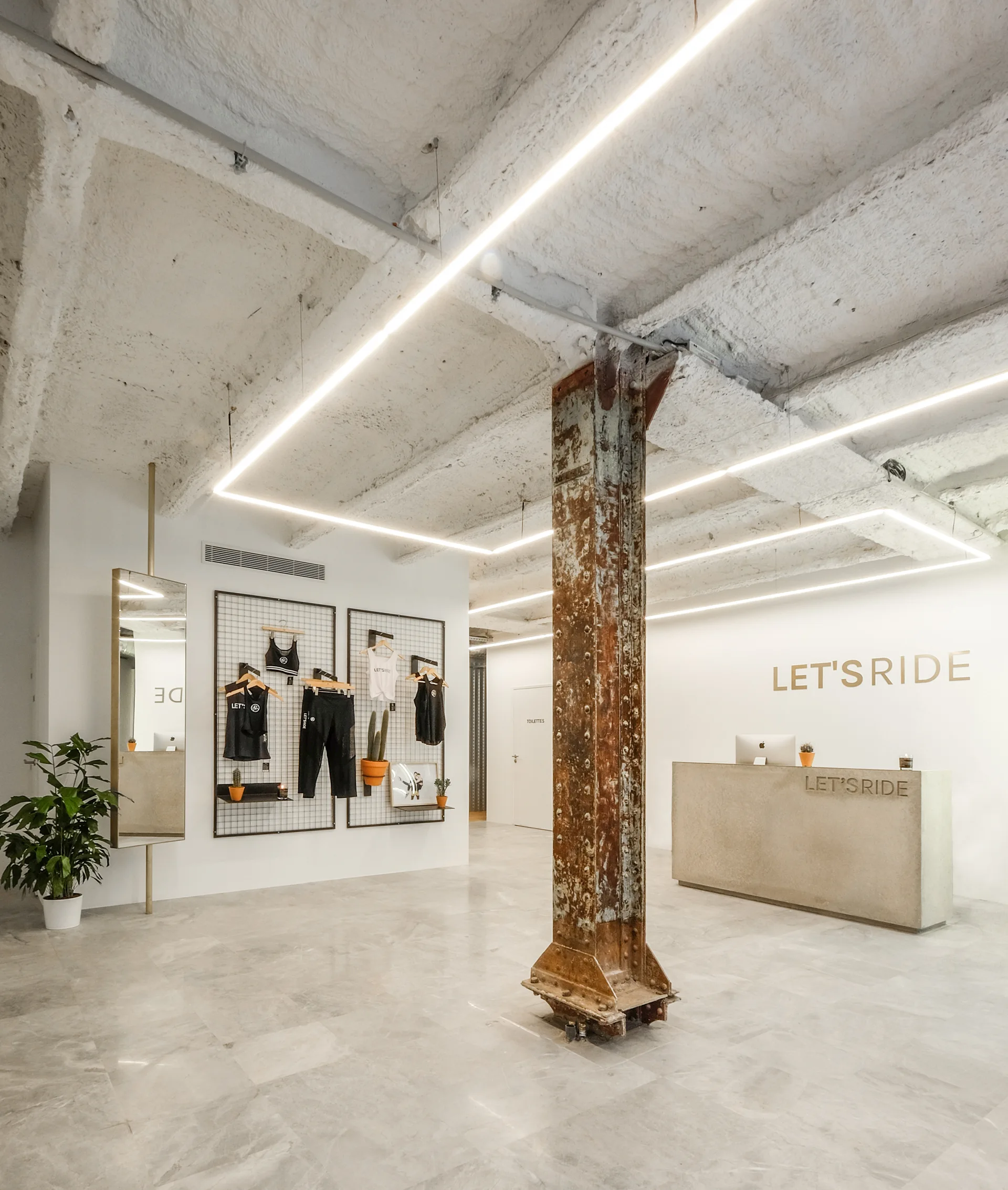 A commercial reception space featuring a central rusted steel column, an exposed concrete ceiling with linear light tracks, and a grey stone floor.
