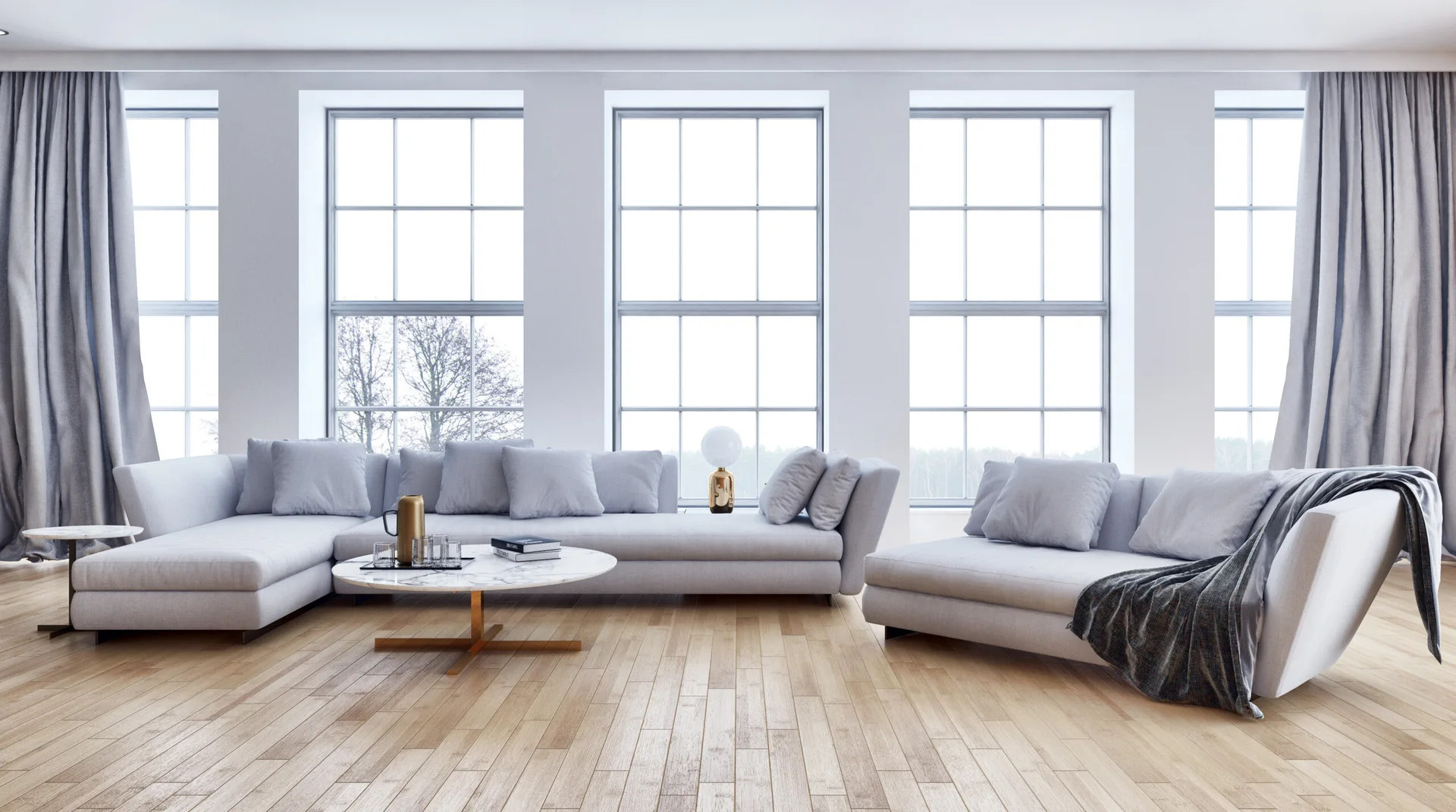 A contemporary living room featuring a light grey sectional sofa, a round marble coffee table, and large multi-pane windows with grey curtains.