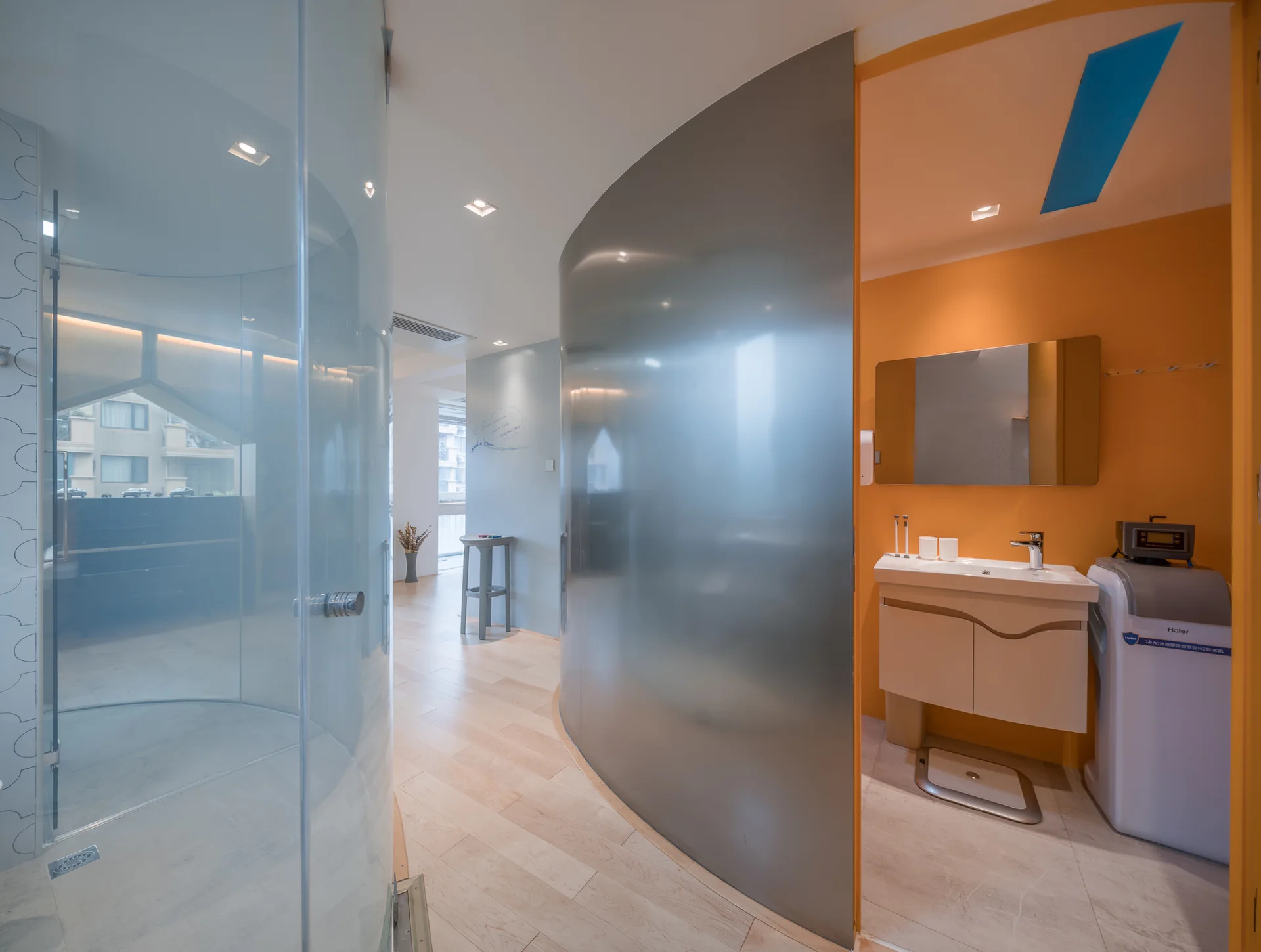 A modern bathroom features a curved metal partition wall, a glass shower enclosure, and a vanity sink set against a bright orange wall.