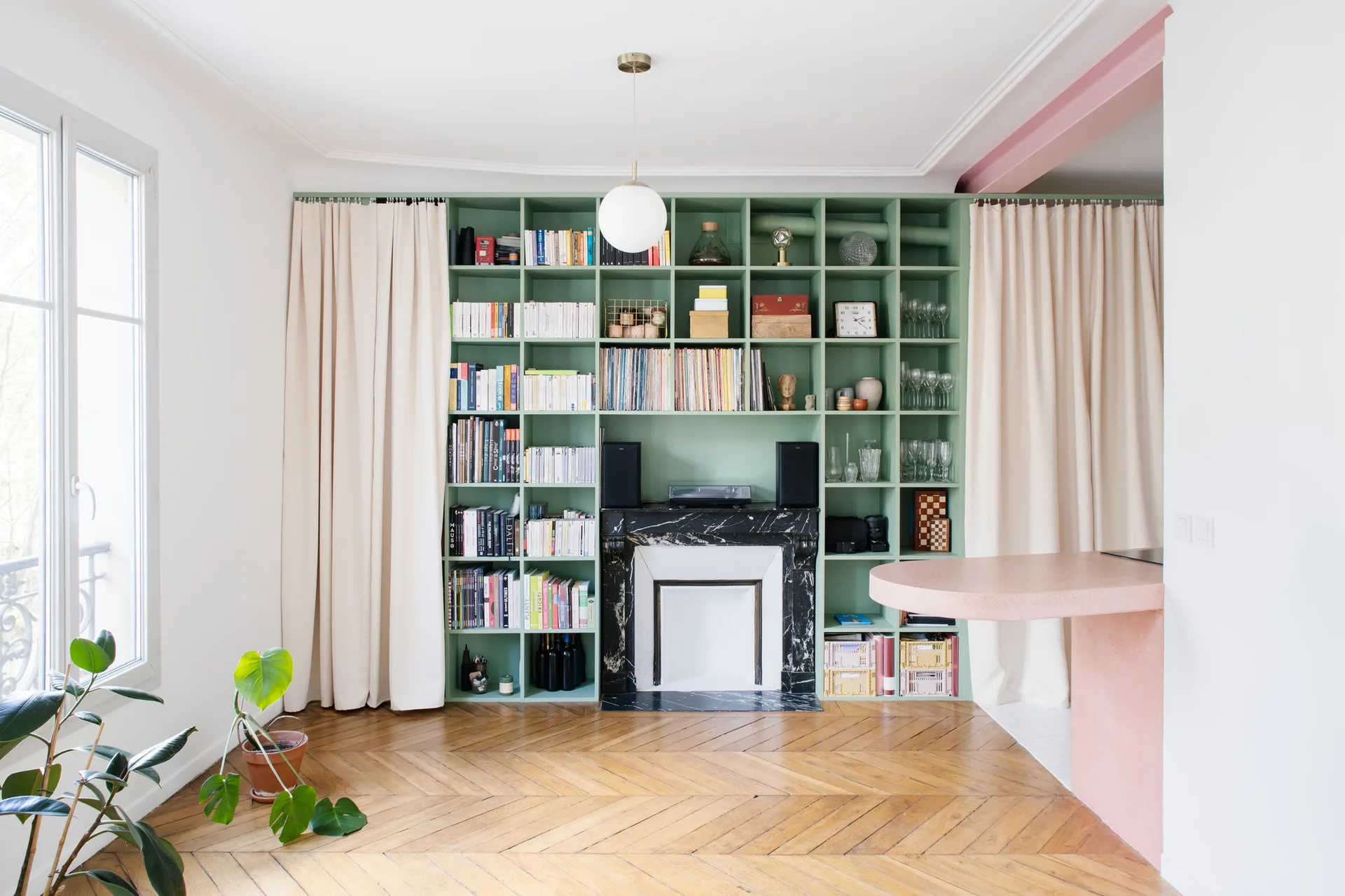 A living room with a sage green floor-to-ceiling bookshelf built around a black marble fireplace, featuring herringbone hardwood floors and off-white curtains.