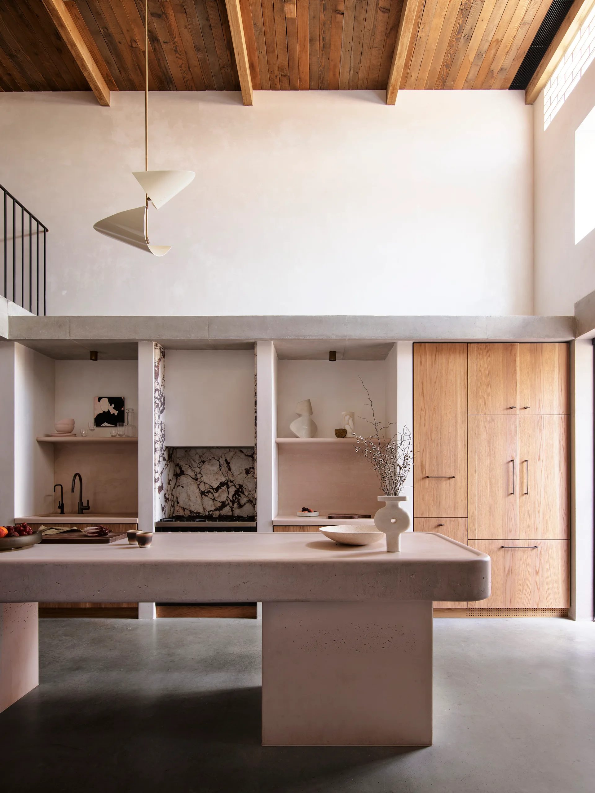 A minimalist kitchen featuring a pale pink concrete island, integrated oak cabinets, and recessed wall alcoves under an exposed wooden ceiling.