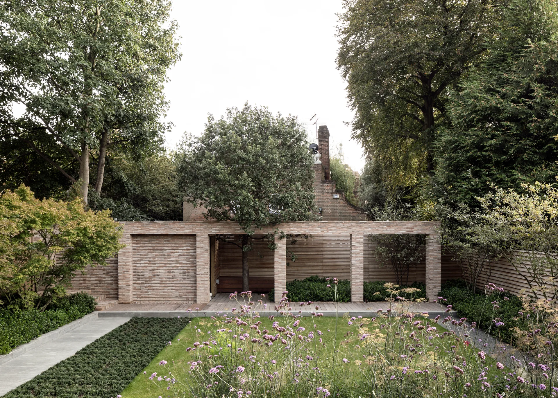 A modern brick pavilion with wood decking overlooks a green lawn and wildflower beds surrounded by mature trees.