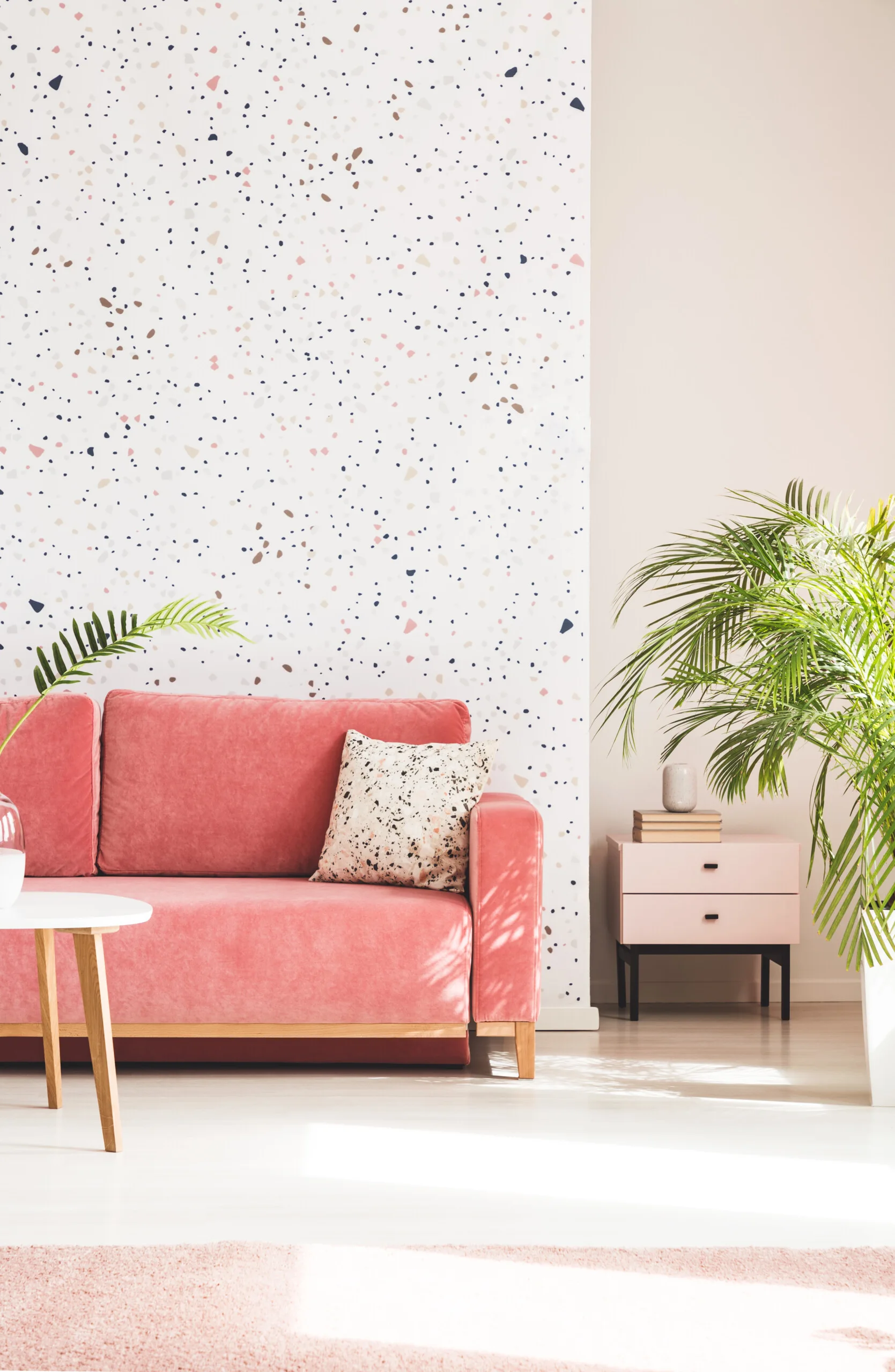 A pink upholstered sofa stands against a terrazzo-patterned accent wall next to a light pink side table and a large green potted palm.