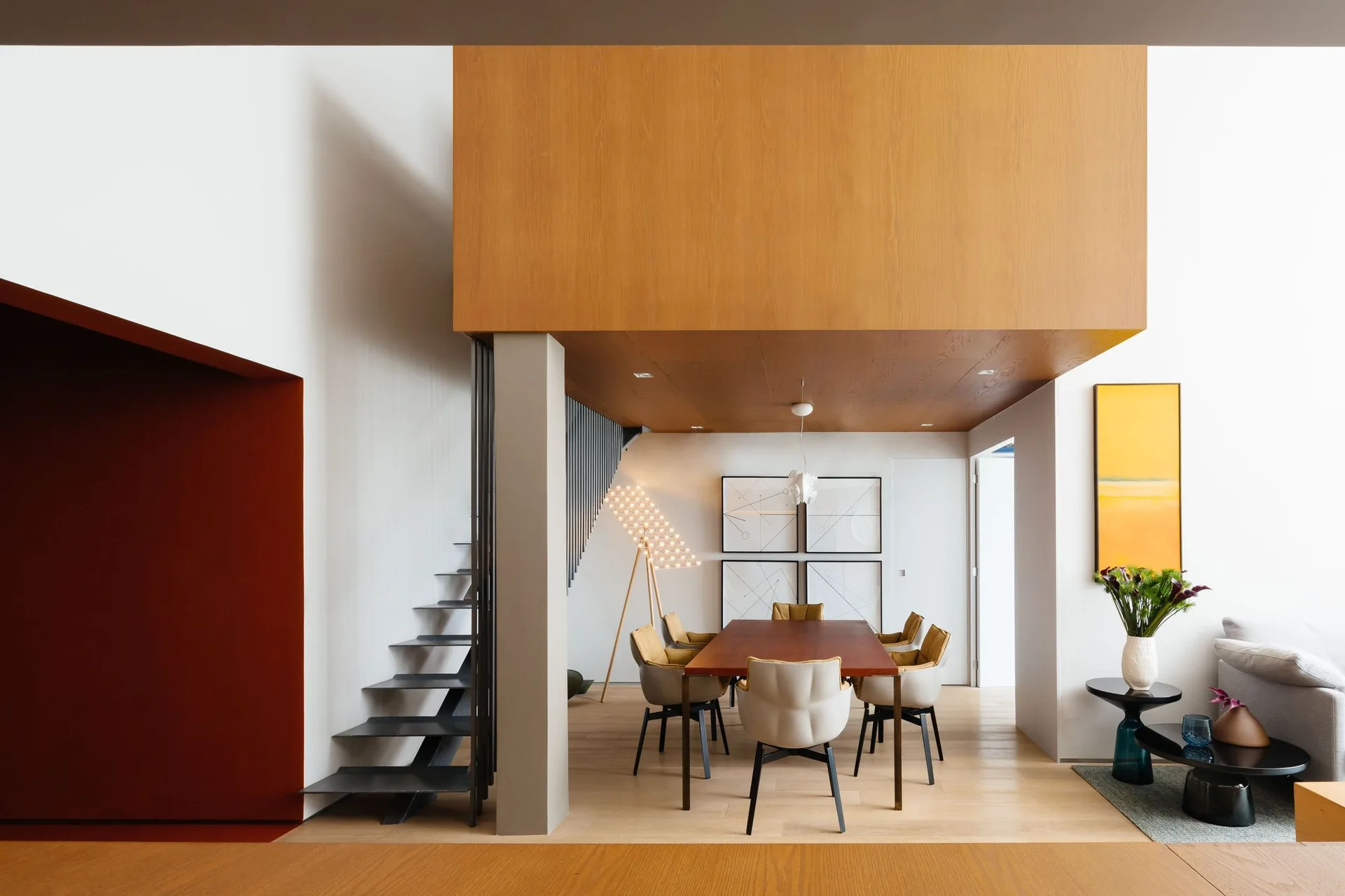 A contemporary dining room features a wood-clad ceiling canopy, a rectangular wooden table with upholstered chairs, and an open metal staircase.