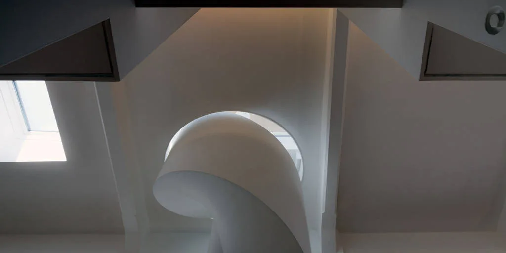 A low-angle view of a minimalist white sculptural spiral staircase leading up toward a ceiling skylight.