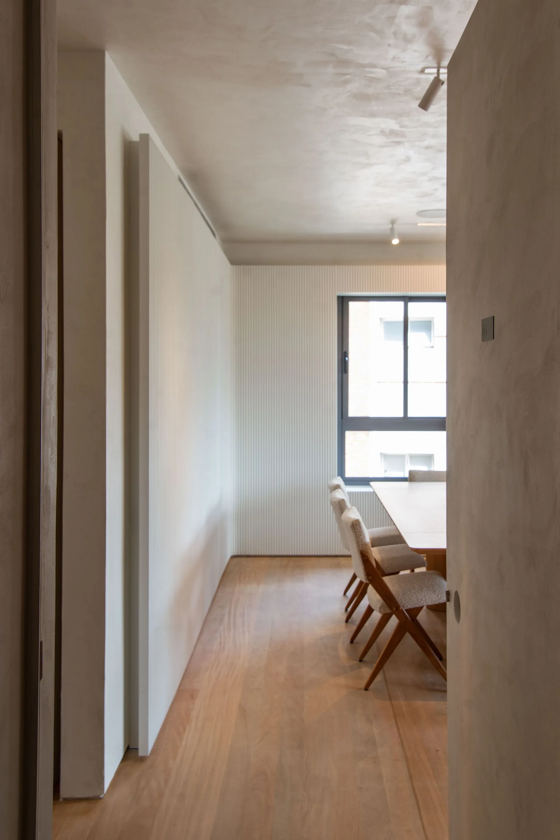 A view looking into a minimalist dining room featuring a wooden table, textured fabric chairs, and a white fluted accent wall around a window.