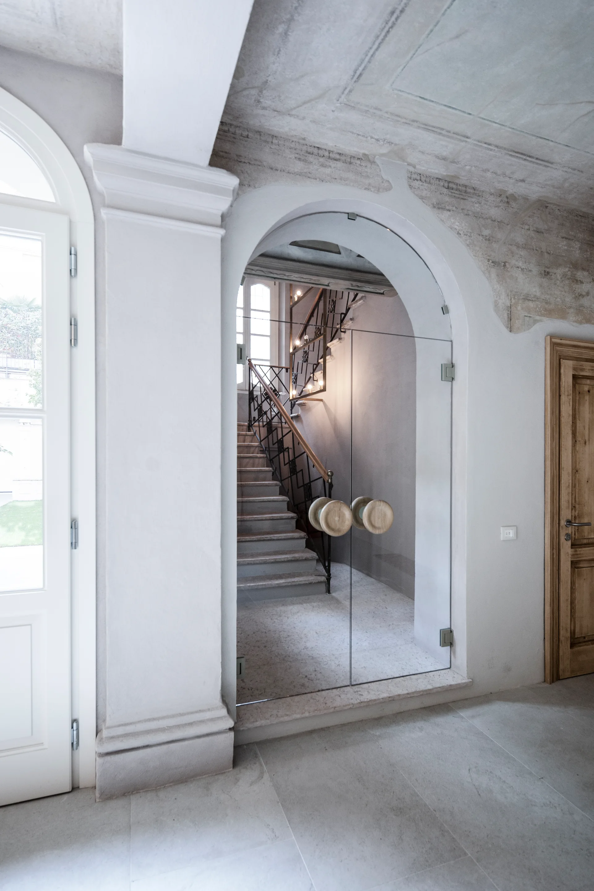 An arched glass door with large round wooden handles opens to a stone staircase with a dark metal banister in a neutral-toned hallway.