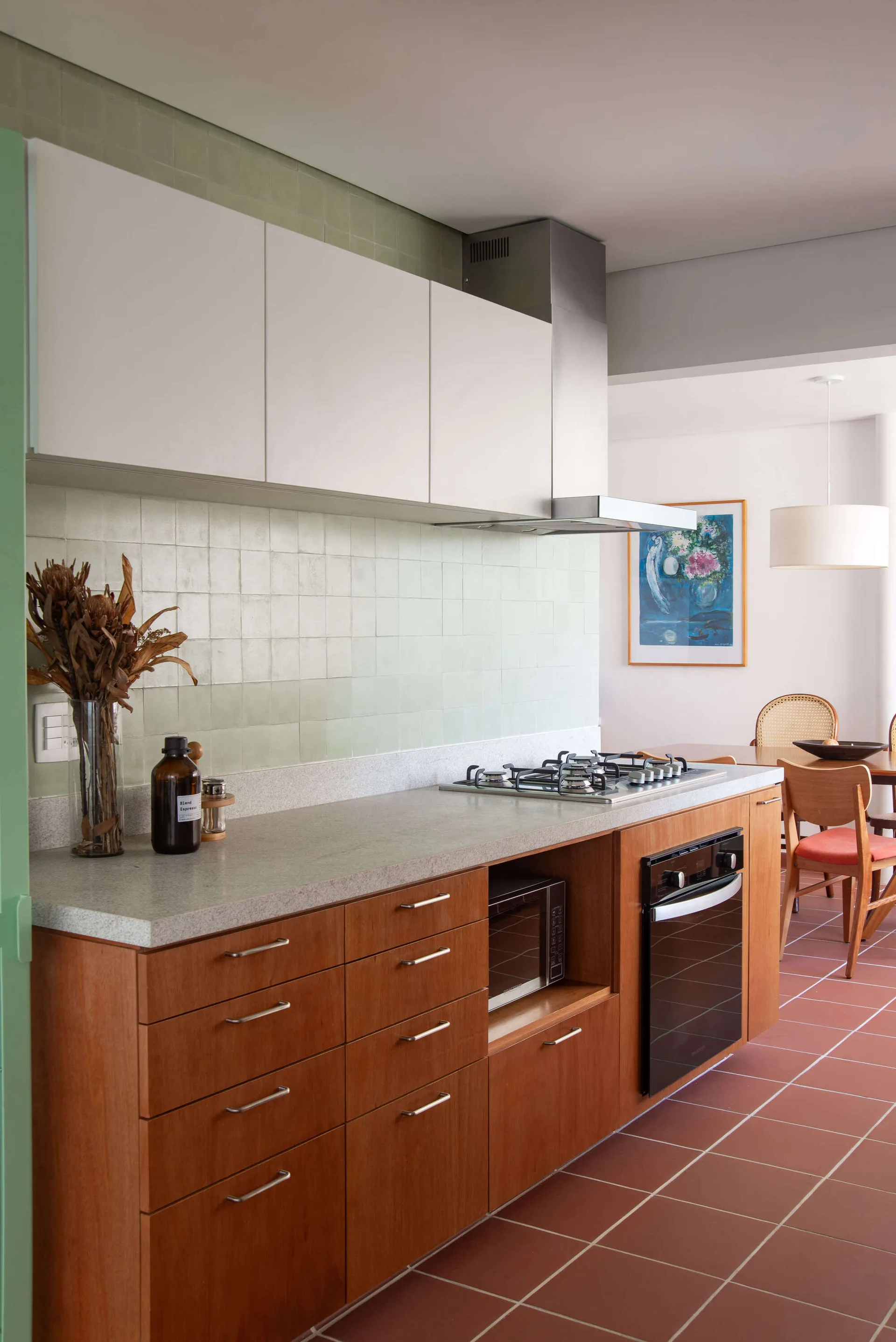 A kitchen featuring warm wood lower cabinets, a light green square-tiled backsplash, and terracotta tile floors leading to a dining area.