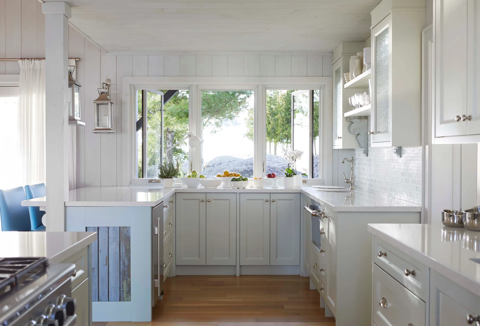 A bright cottage kitchen features pale blue cabinetry, white countertops, vertical wood wall paneling, and a large triple window overlooking trees.