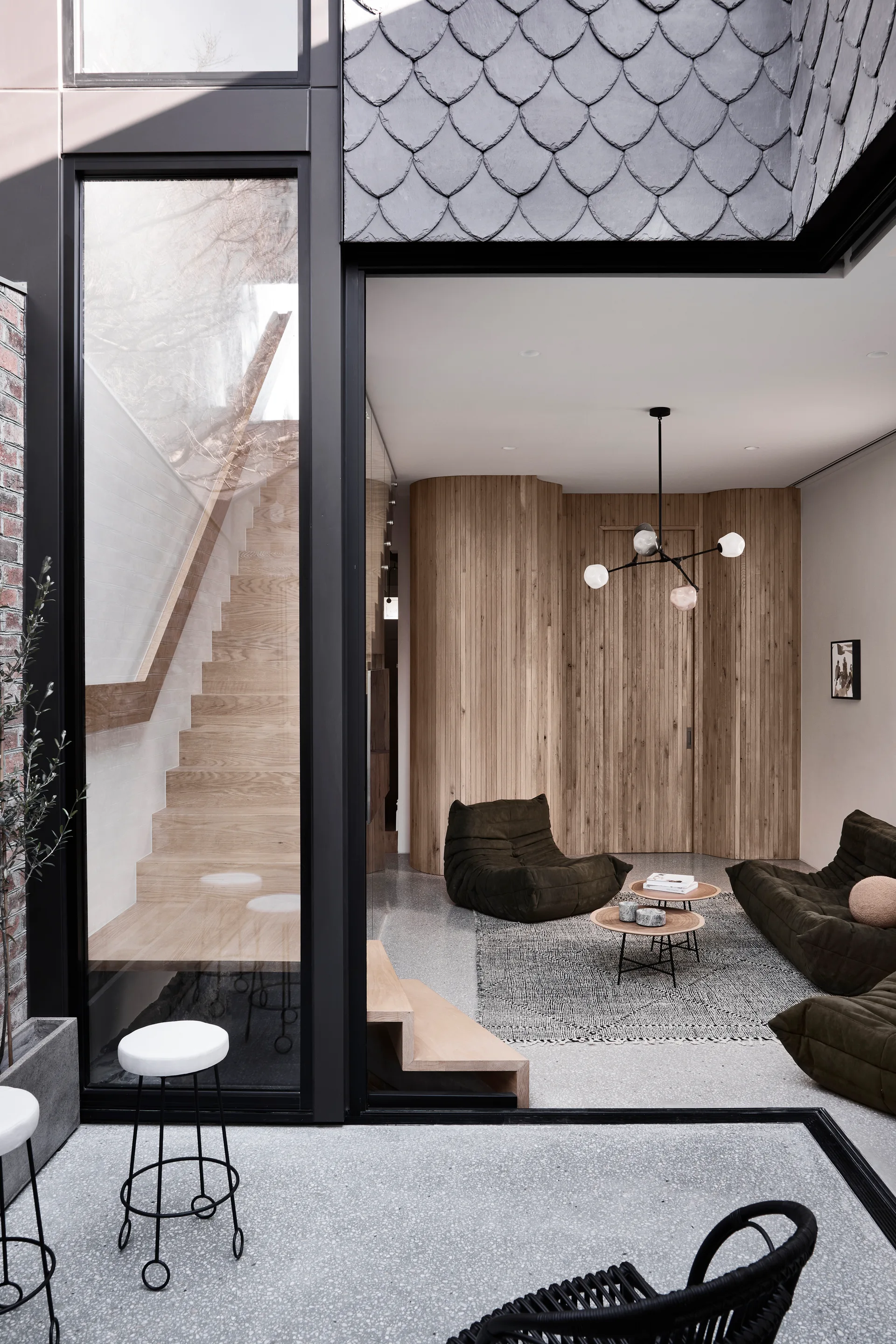 A modern living room features a curved vertical wood slat wall, dark green modular sofas, a textured rug, and an adjacent glass-enclosed oak staircase.