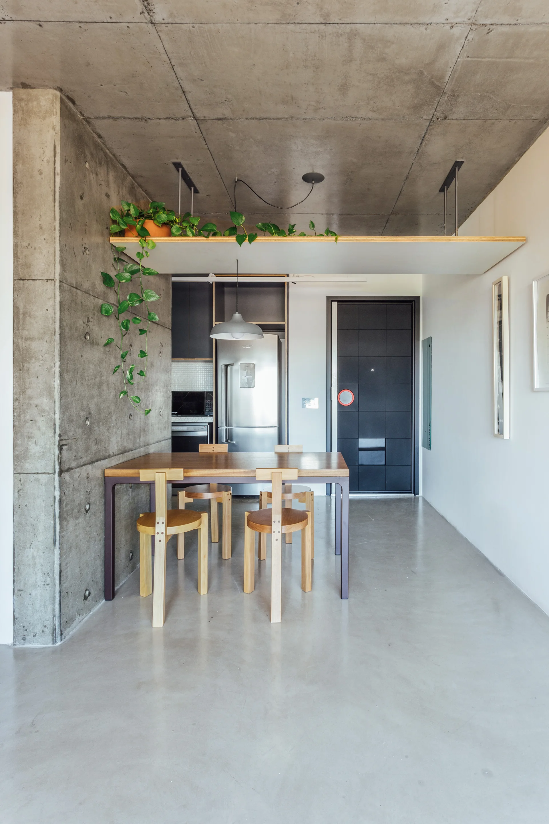An industrial dining area features a wooden table and chairs beneath an exposed concrete ceiling with a suspended shelf holding a trailing plant.