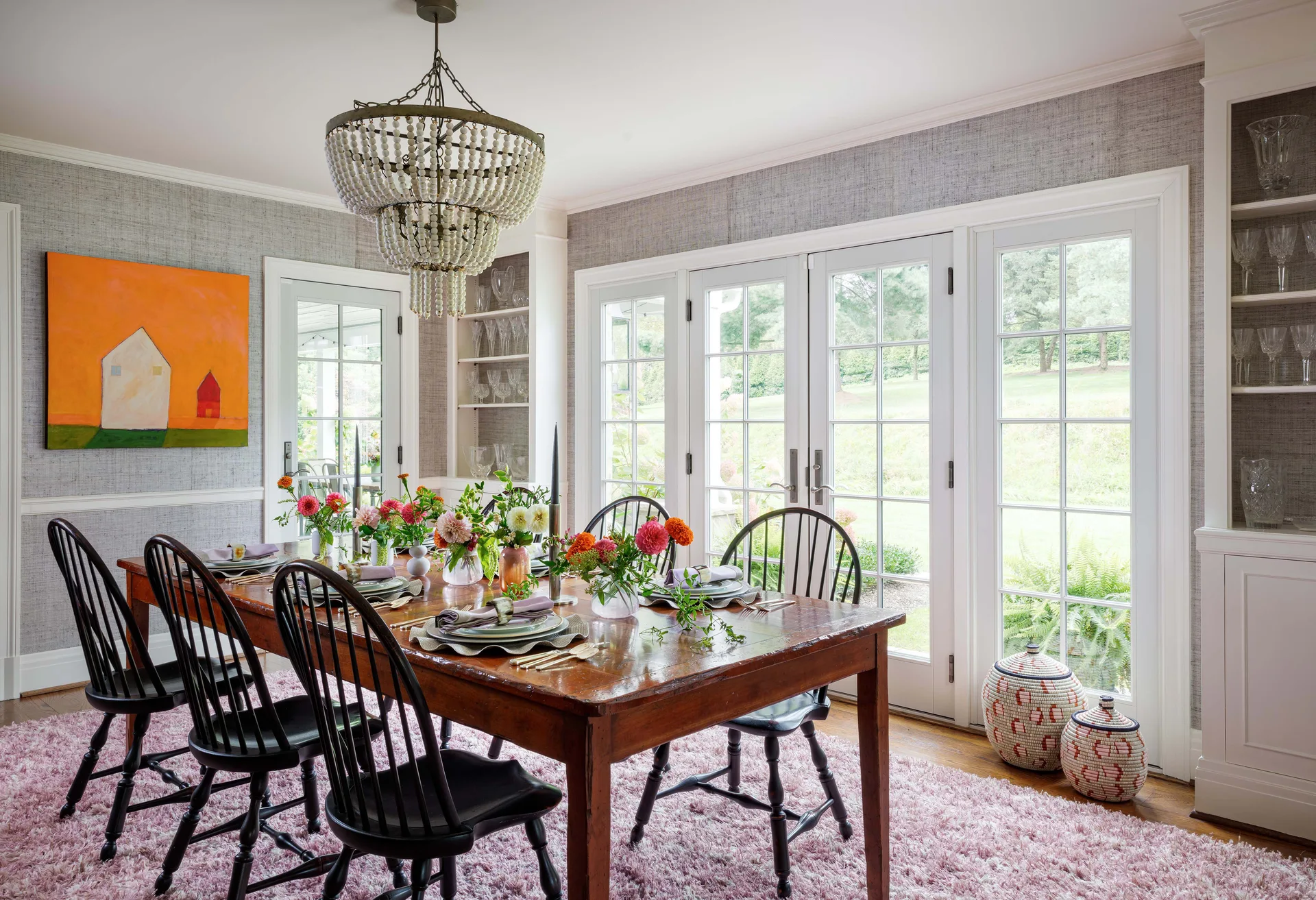 A wooden dining table with black spindle chairs sits on a pink shag rug in a room with grey grasscloth wallpaper and French doors.