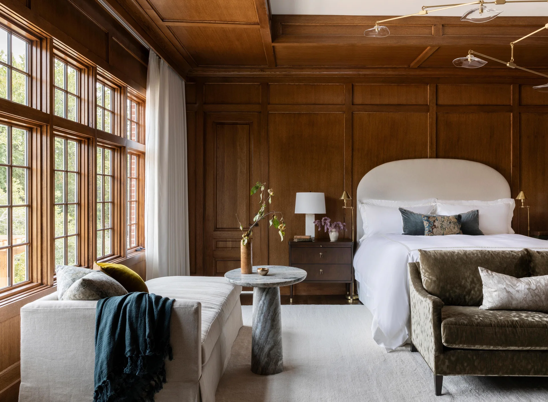 A bedroom featuring dark wood-paneled walls and ceiling, a cream upholstered bed, a matching chaise longue, and a round stone pedestal table.