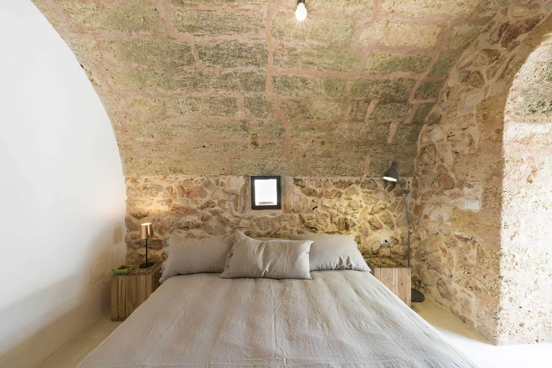 A bed with neutral linen sheets is positioned against a rustic stone wall under a barrel-vaulted stone ceiling.