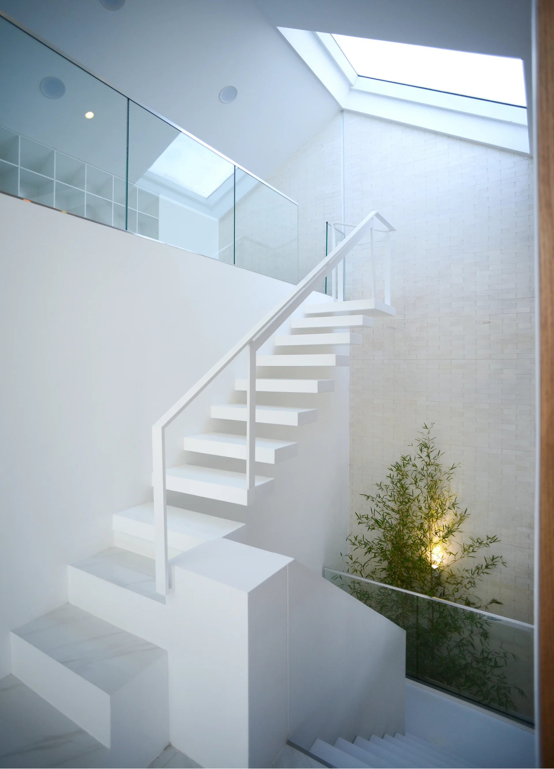 A minimalist white floating staircase with glass railings ascends toward an overhead skylight next to a textured white stone wall.
