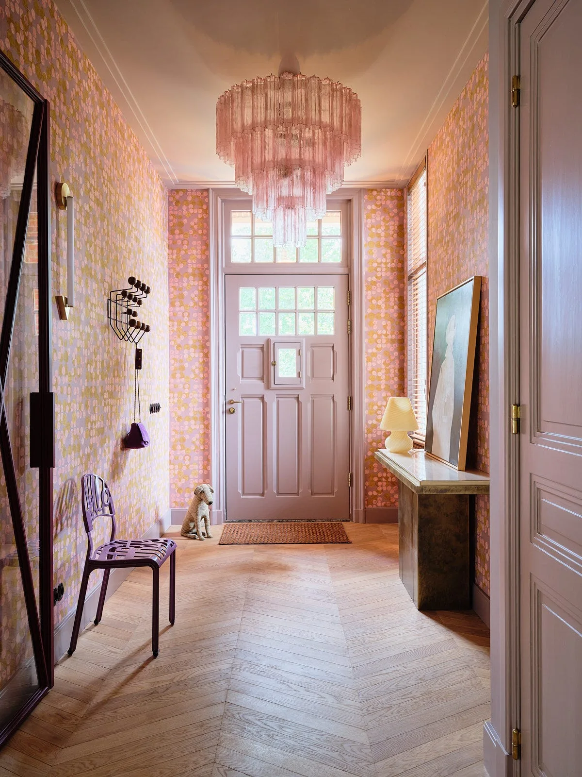 An entryway hallway features patterned pink wallpaper, chevron wood flooring, a tiered pink fringe chandelier, and a lavender front door.