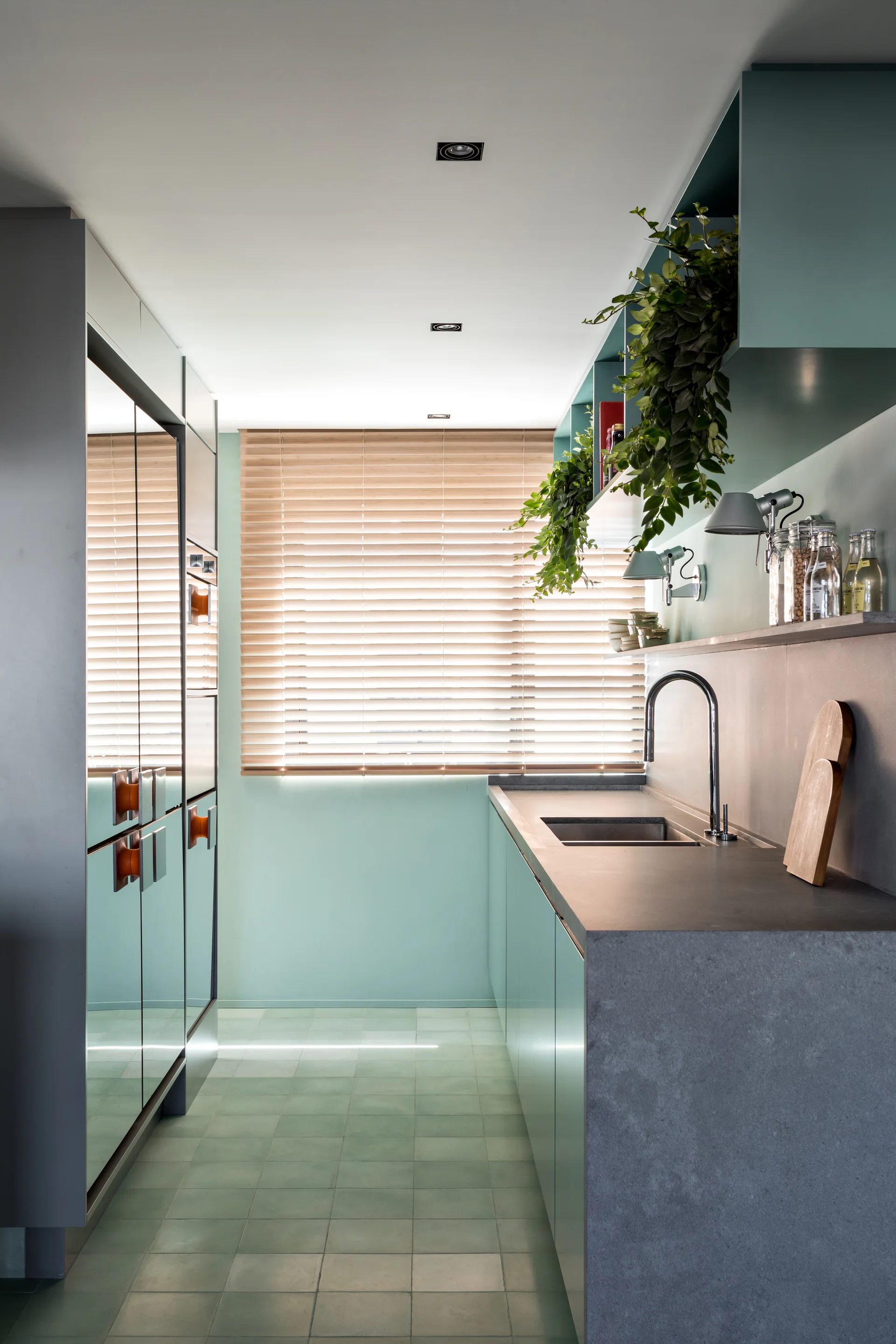 A narrow galley kitchen features mint green cabinets, a grey concrete countertop, mirrored cabinet doors, and wooden venetian blinds on the window.