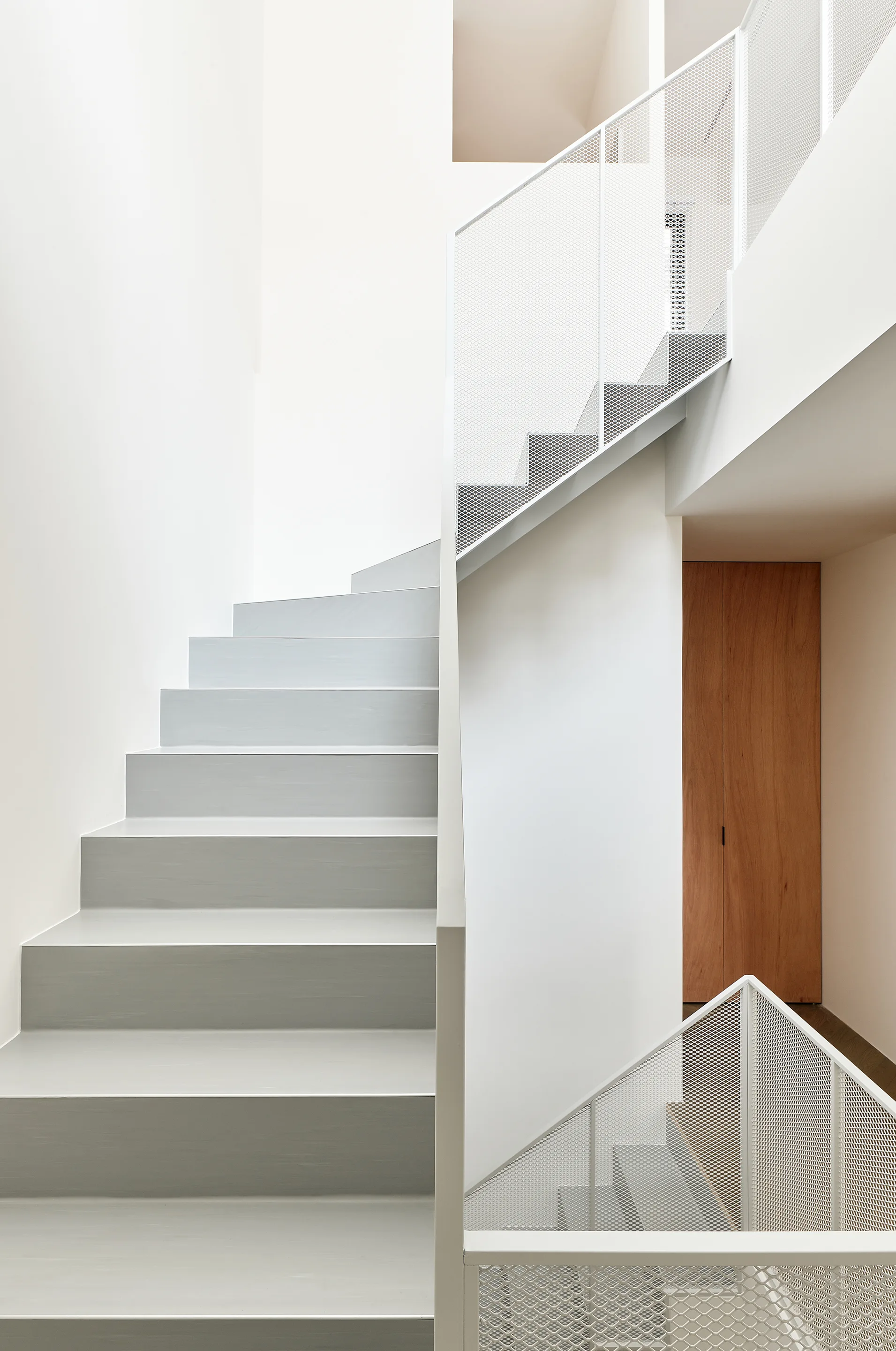 A minimalist modern staircase features light gray steps and white metal mesh railings against white walls with a natural wood door in the background.