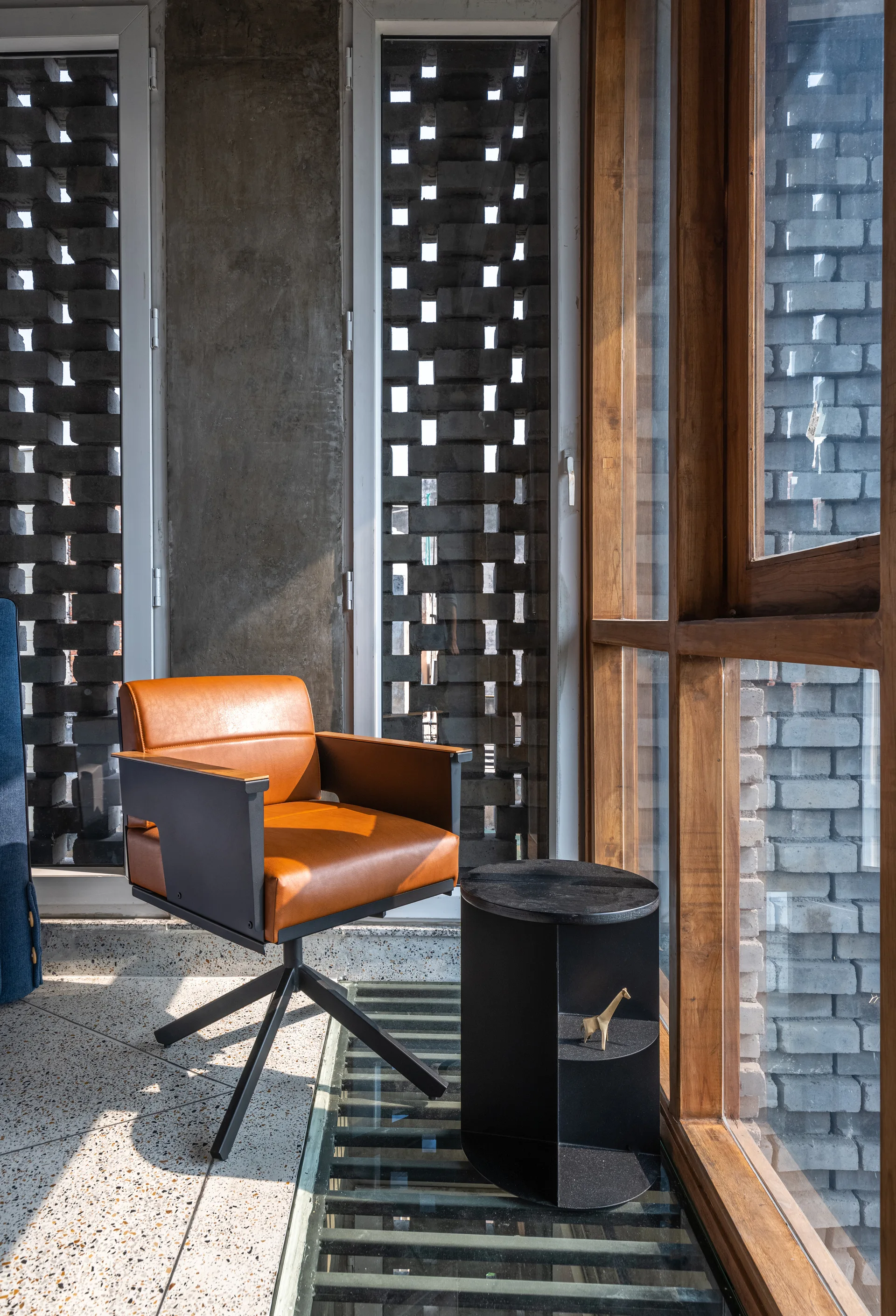 A tan leather armchair and a black side table sit on a polished terrazzo floor next to a large window showing a patterned brick screen.