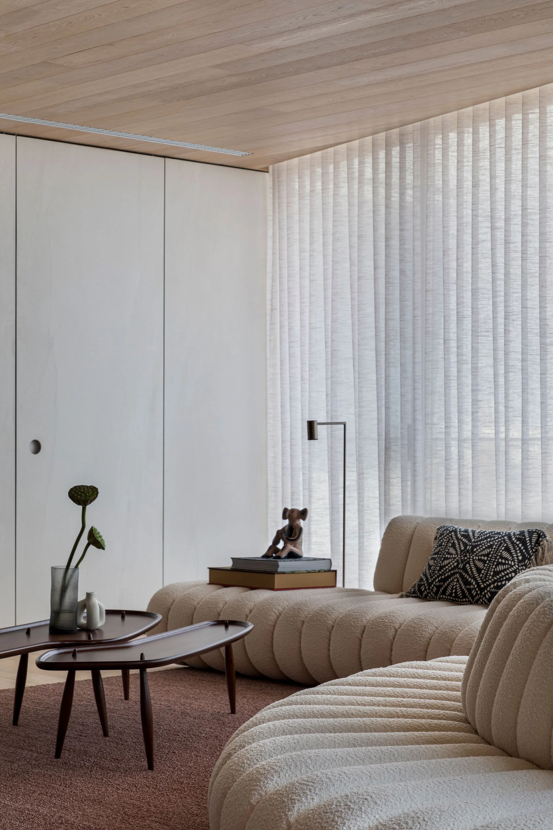 A minimalist living room featuring a cream ribbed sofa, nested wooden coffee tables, and sheer white curtains under a wood plank ceiling.