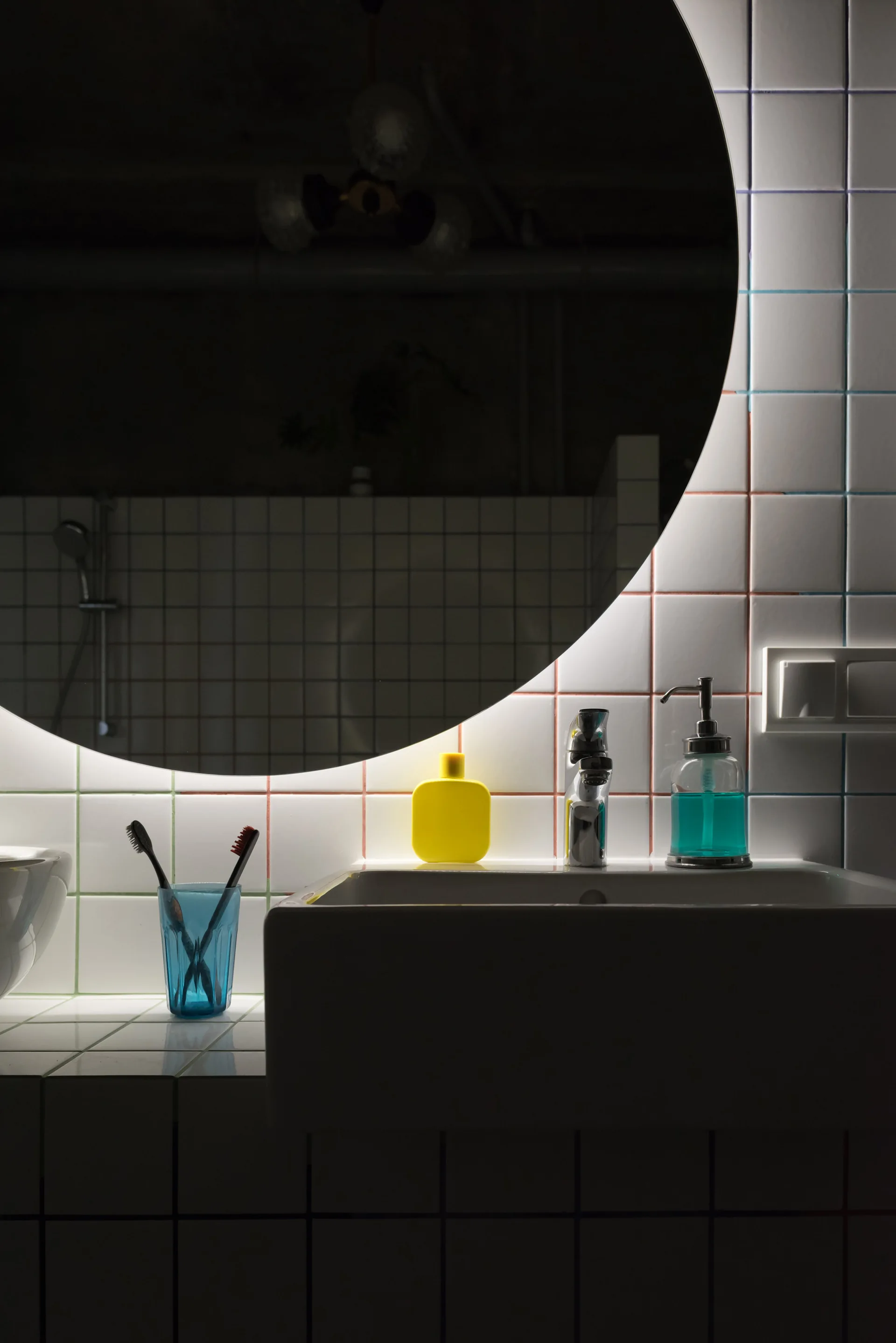 A round backlit mirror hangs above a white sink against a white grid-tiled wall with colorful grout lines.