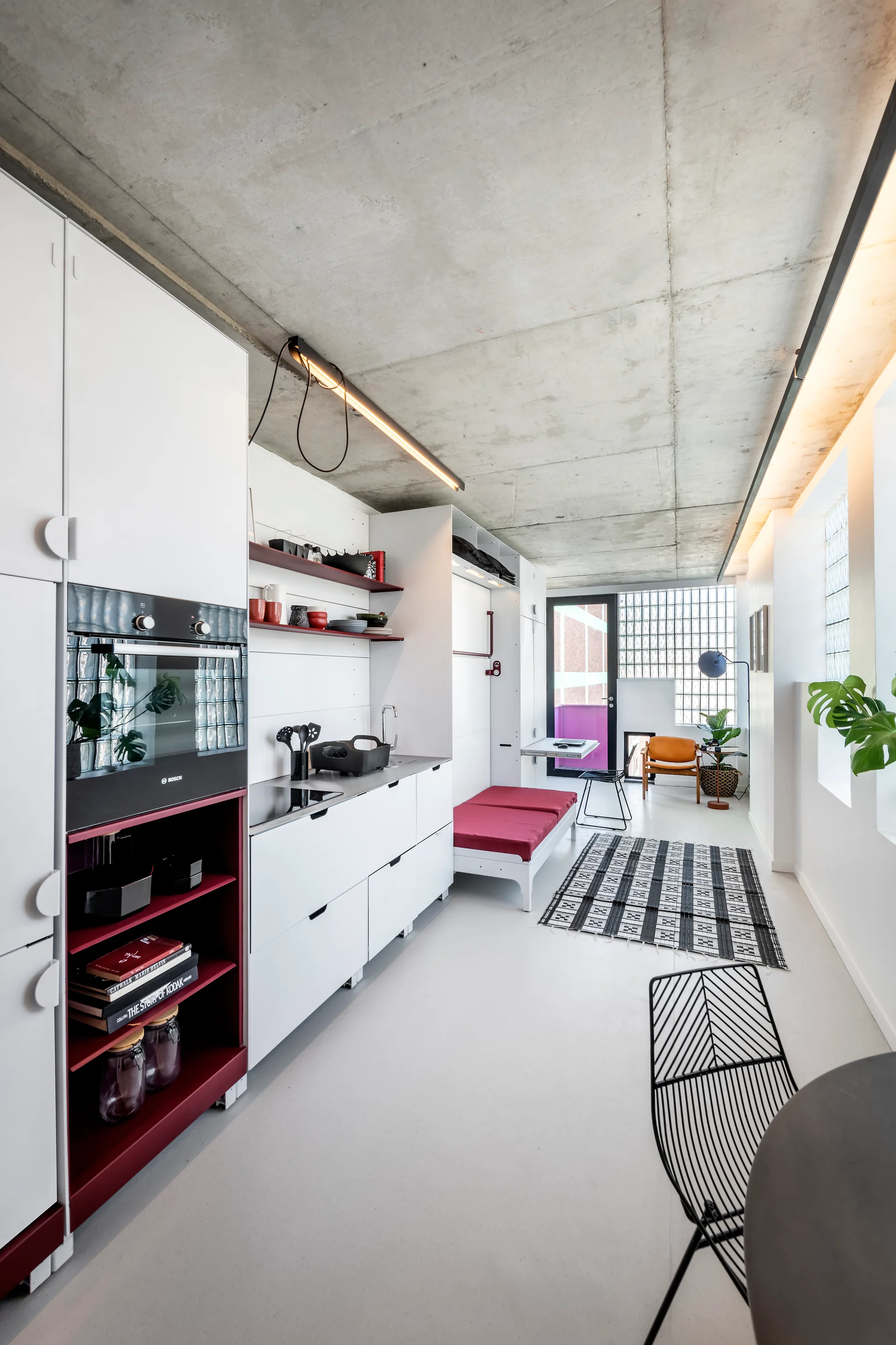 A modern studio apartment features flat-front white kitchen cabinets with deep red accents beneath an exposed concrete ceiling with industrial lighting.