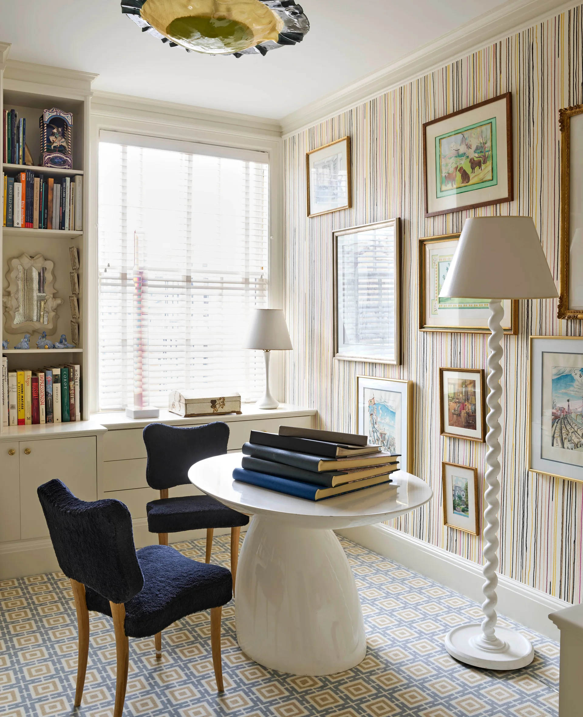 A bright library room features a white pedestal table, two dark blue chairs, built-in bookshelves, and framed art against striped wallpaper.