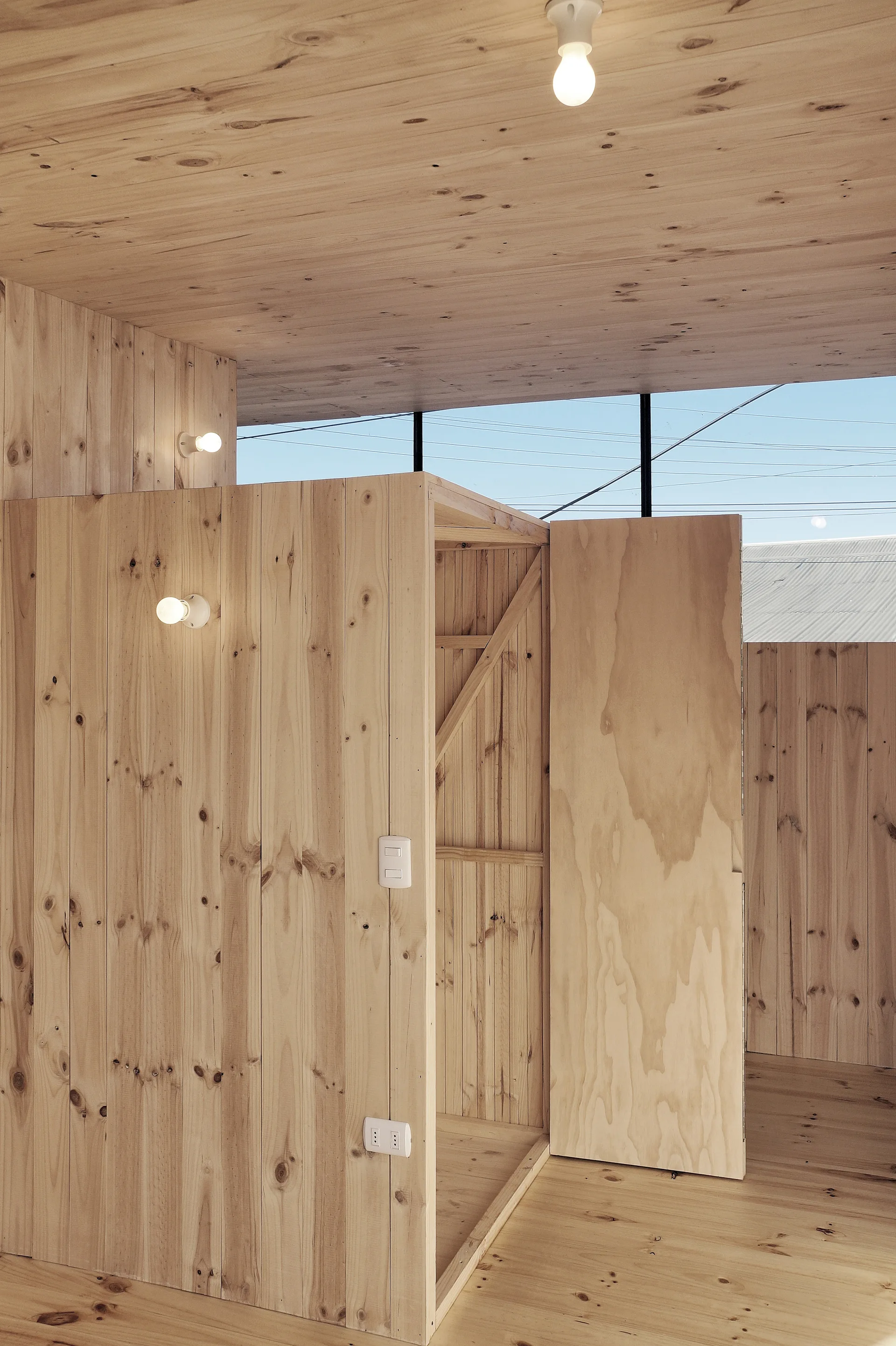 A minimalist room interior featuring light-toned wood panels on the floors, walls, and ceiling, with simple exposed bulb light fixtures.