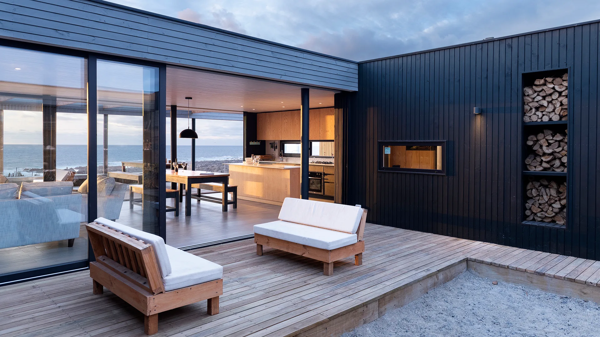 A modern dark-clad house with a wooden patio, outdoor benches, sliding glass doors, and a built-in vertical firewood storage niche.